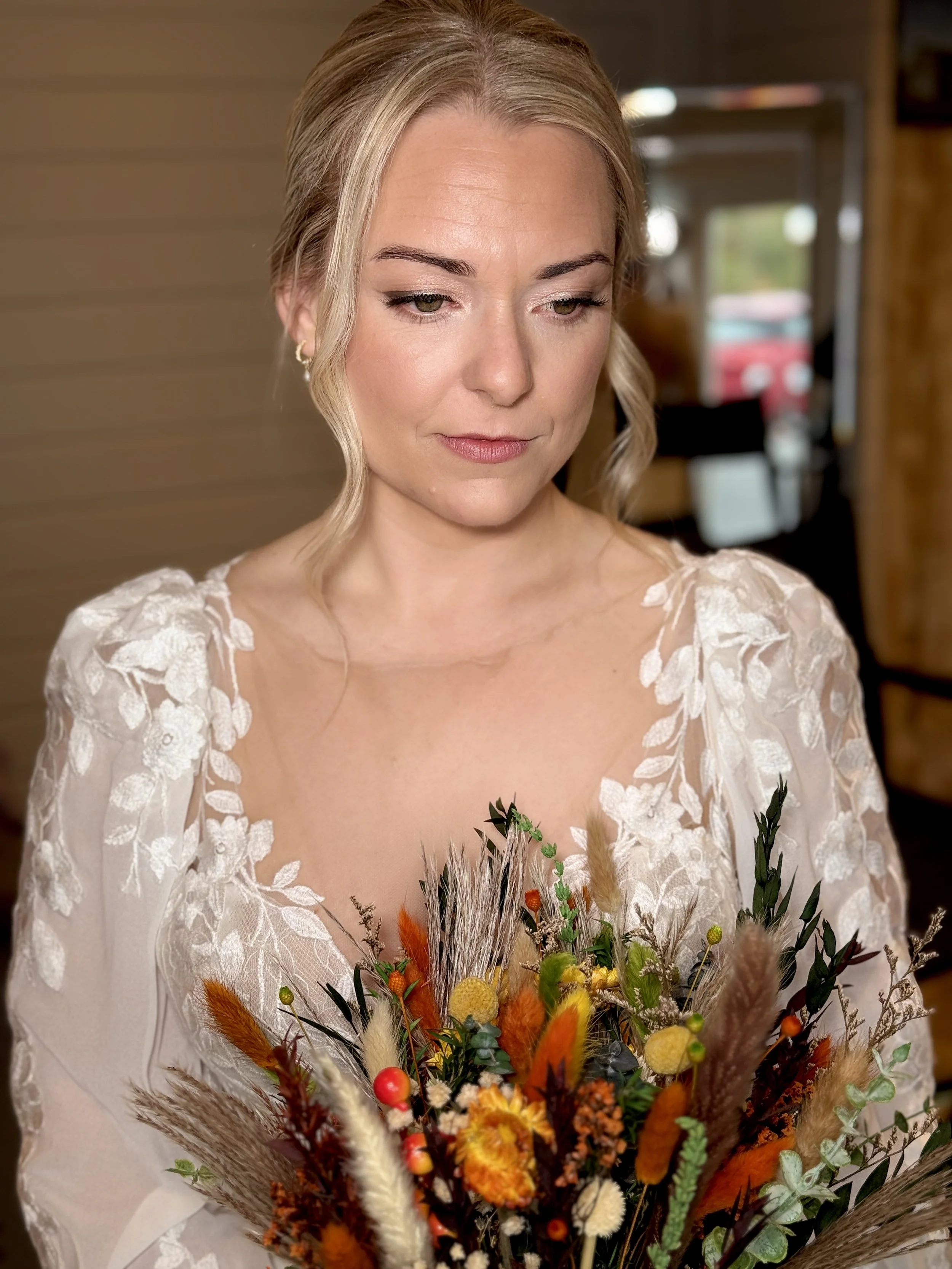 A woman in a white lace dress holding a colorful bouquet of flowers, looking down softly.
