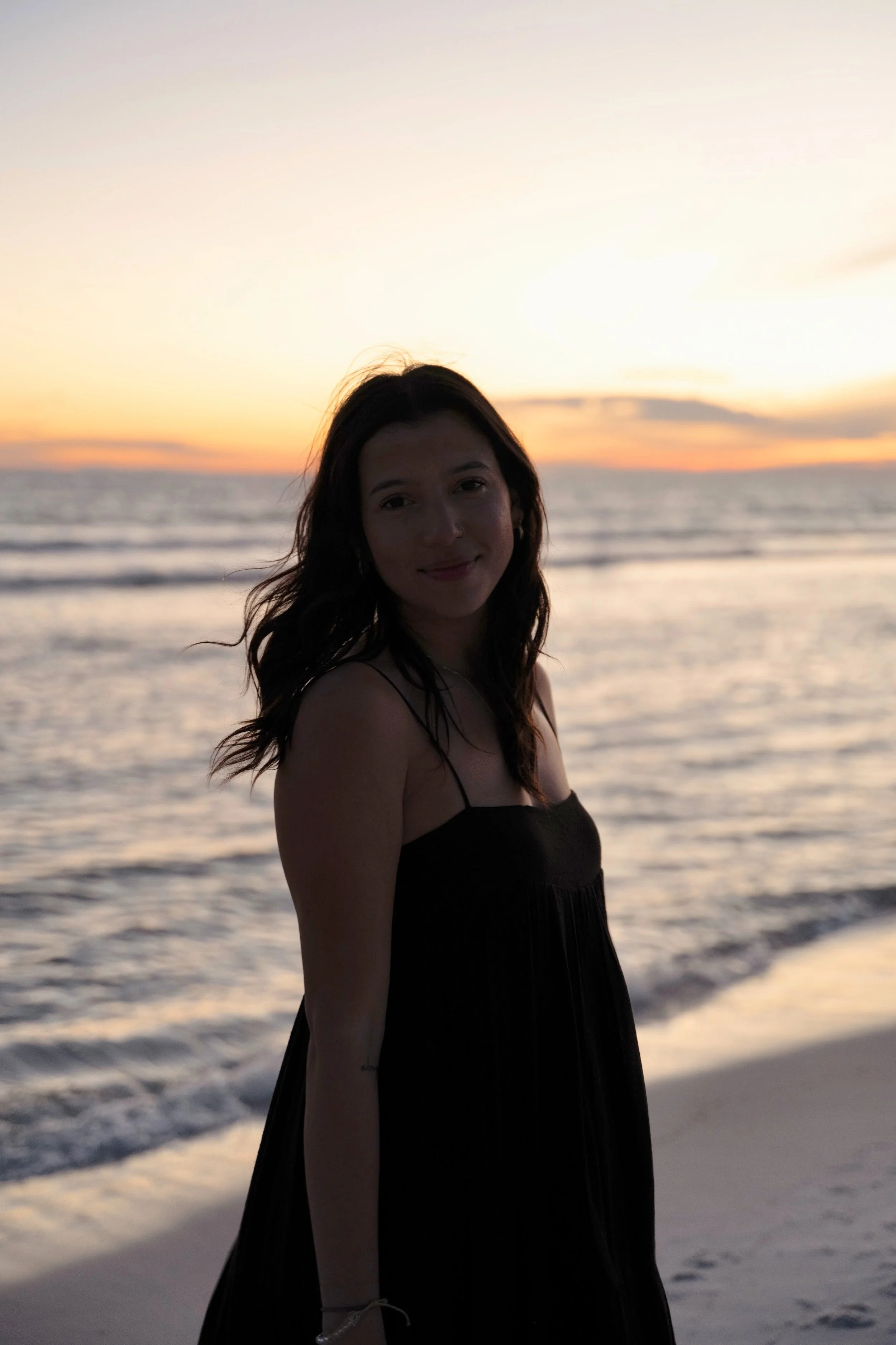 A woman standing on the beach at sunset, with the ocean and sky in the background, wearing a black dress.