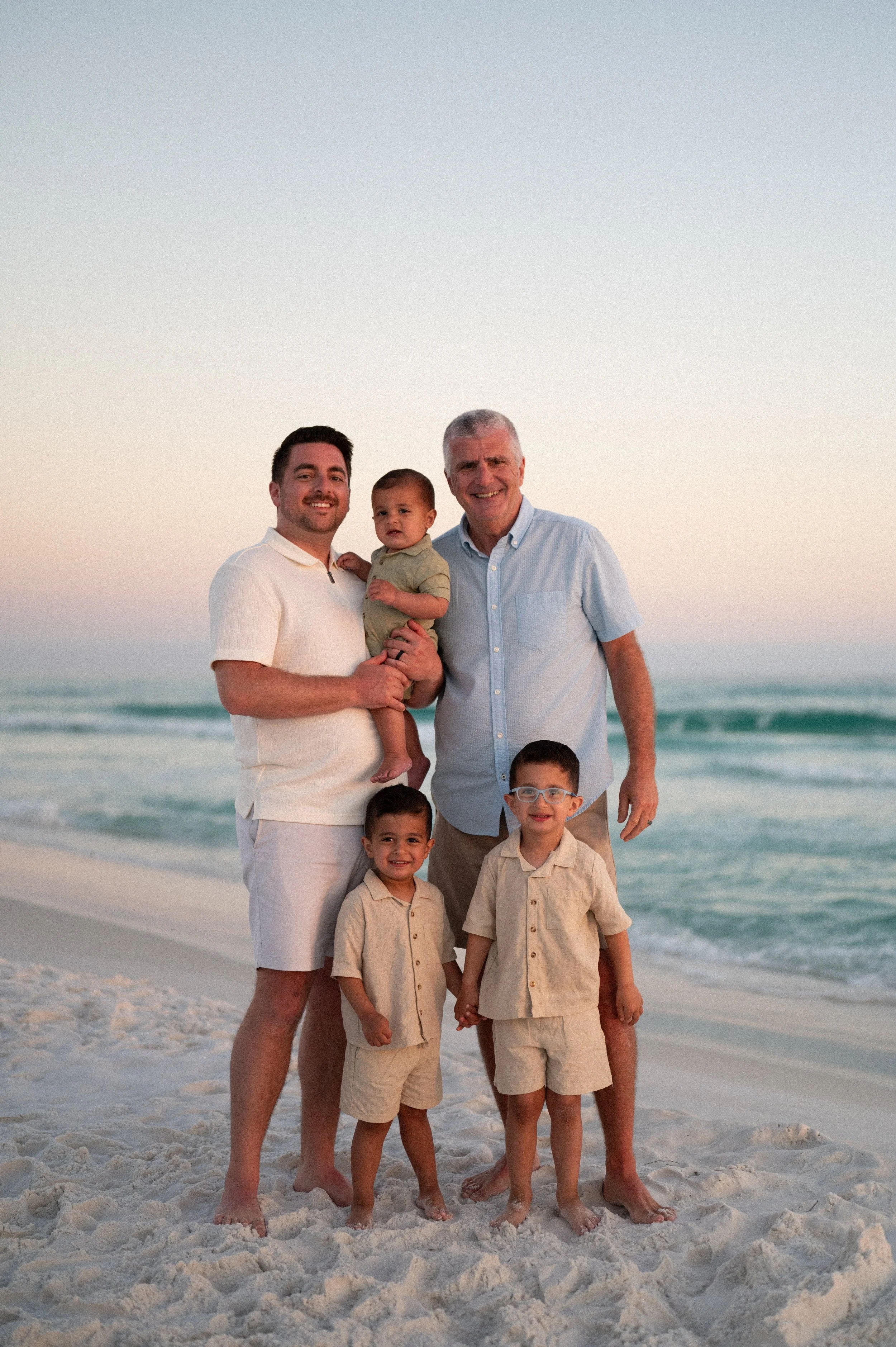 A wedding cake decorated with pink and white icing, topped with a bride and groom figurine, placed on a table surrounded by various arrangements of flowers, with glasses of wine, plates, forks, and a calendar on the table, against a wallpapered background with city and countryside scenes.