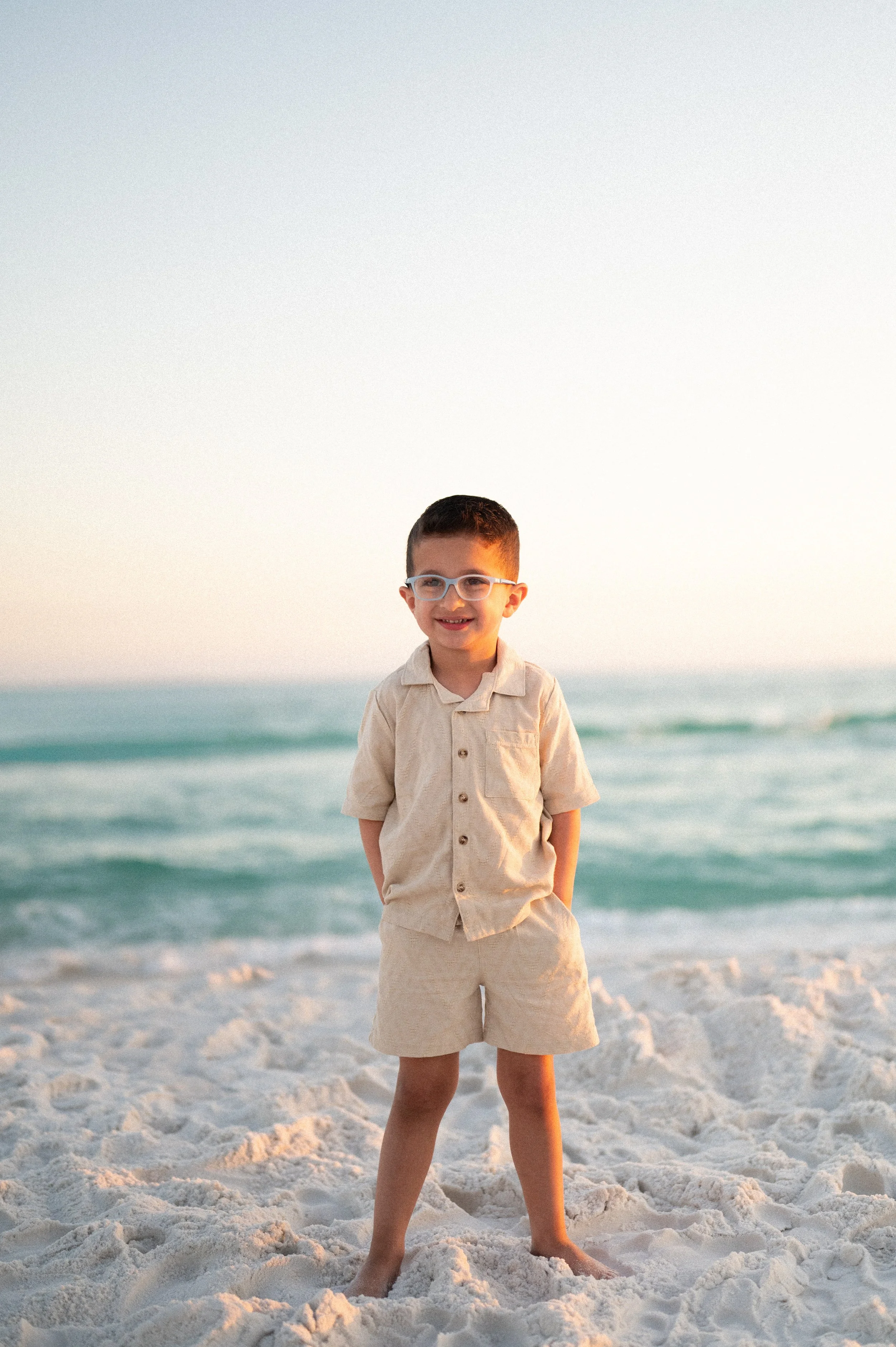 Elegant outdoor wedding reception setup with a long white table decorated with green foliage and white flowers, surrounded by white chairs and lanterns, under hanging paper lanterns and string lights in a lush green setting.