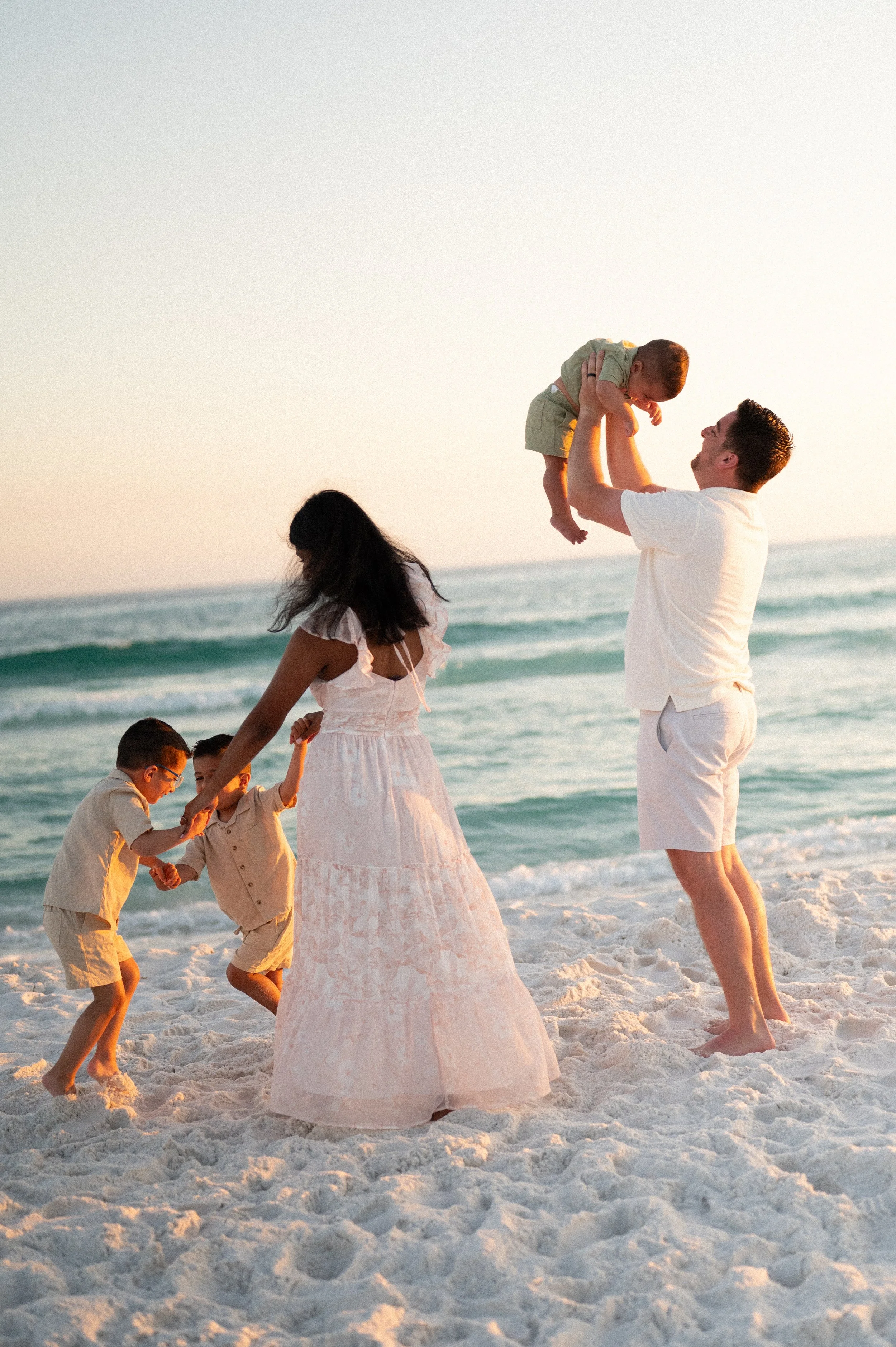 A newlywed couple walks arm in arm through a celebratory outdoor wedding reception, surrounded by guests throwing paper confetti, with a wooden arch and lush green landscape in the background.
