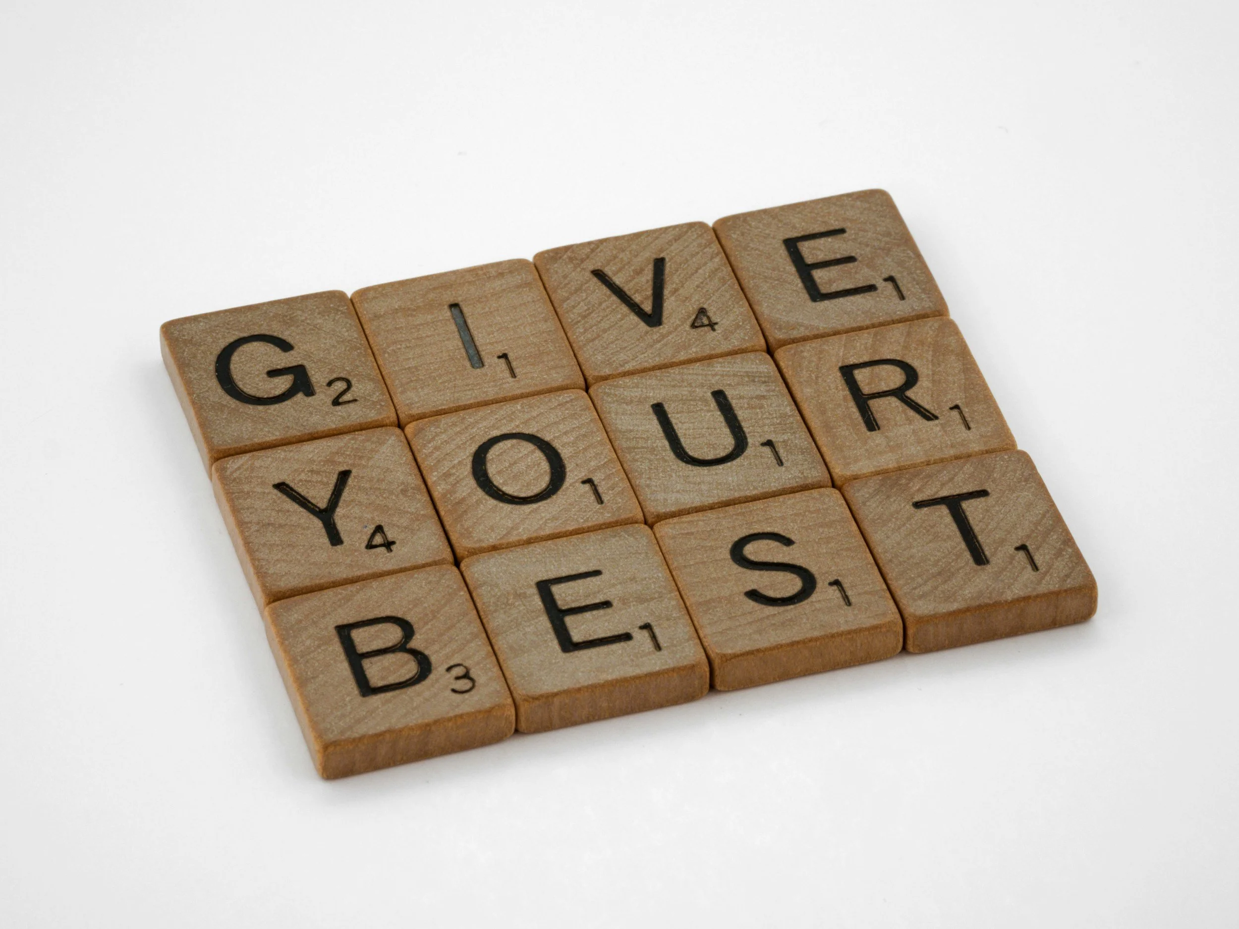 Close-up of wooden Scrabble tiles spelling the word "GIVE" on a white background.