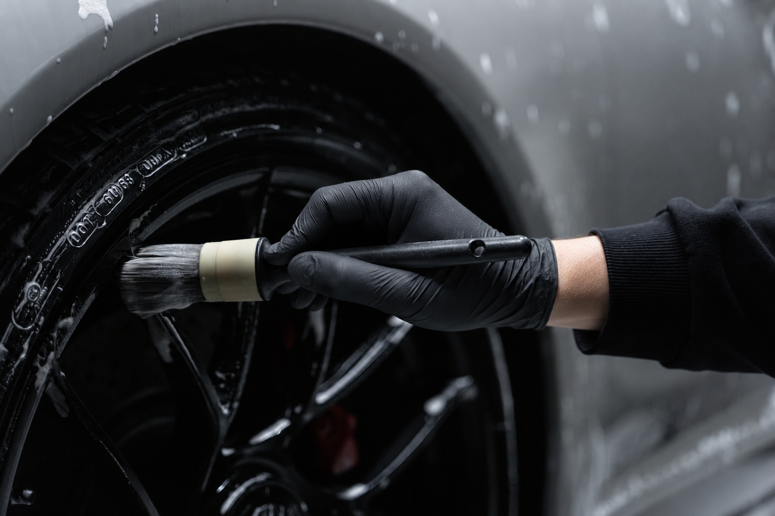 A person wearing black gloves is cleaning a black car wheel with a brush and soap.