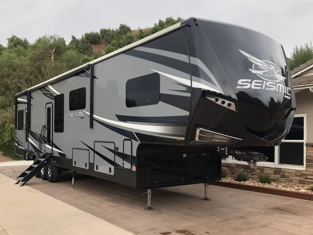 A large black and gray Seismic brand fifth wheel RV parked on a concrete driveway in front of a house with a stone exterior and a garden. The RV has multiple windows and a set of stairs leading to the door.