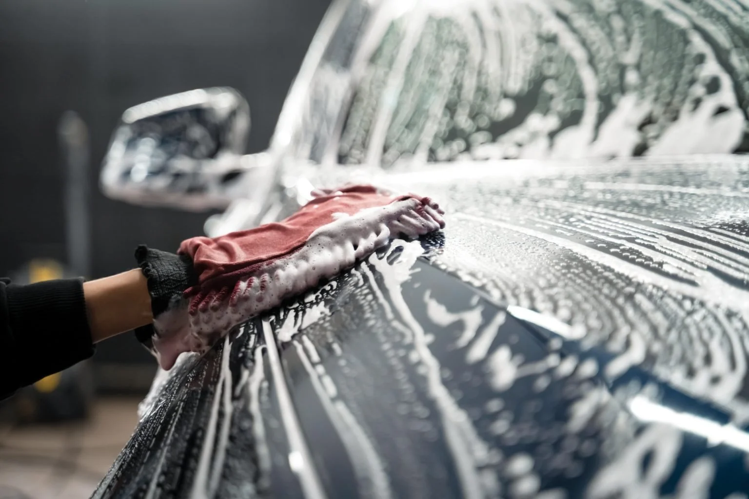 Person washing a dark-colored car with a sponge covered in soap, creating foam on the surface.