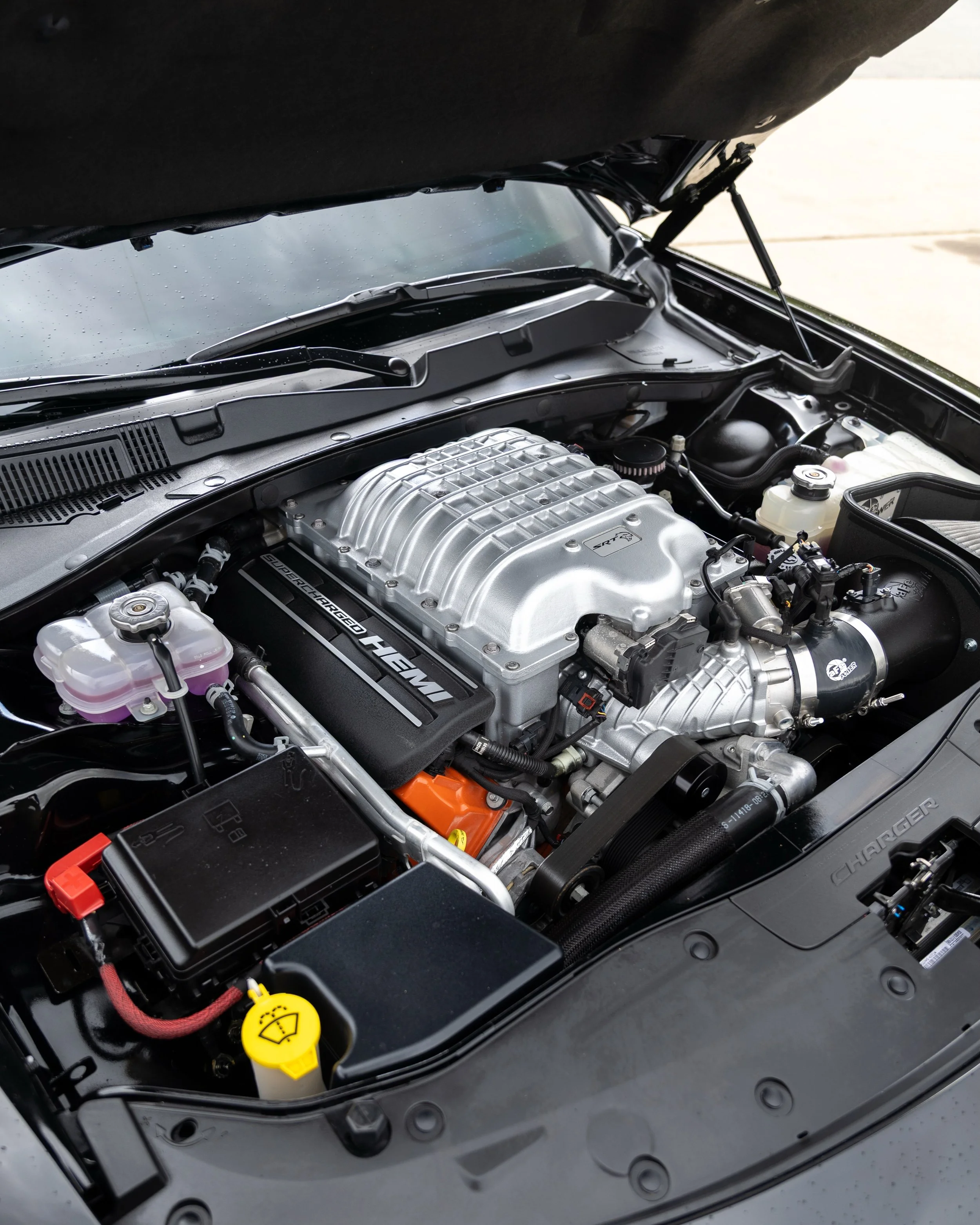 Car hood open revealing engine with supercharged HEMI label, power steering fluid reservoir, and windshield washer fluid reservoir with yellow cap, on a concrete driveway.