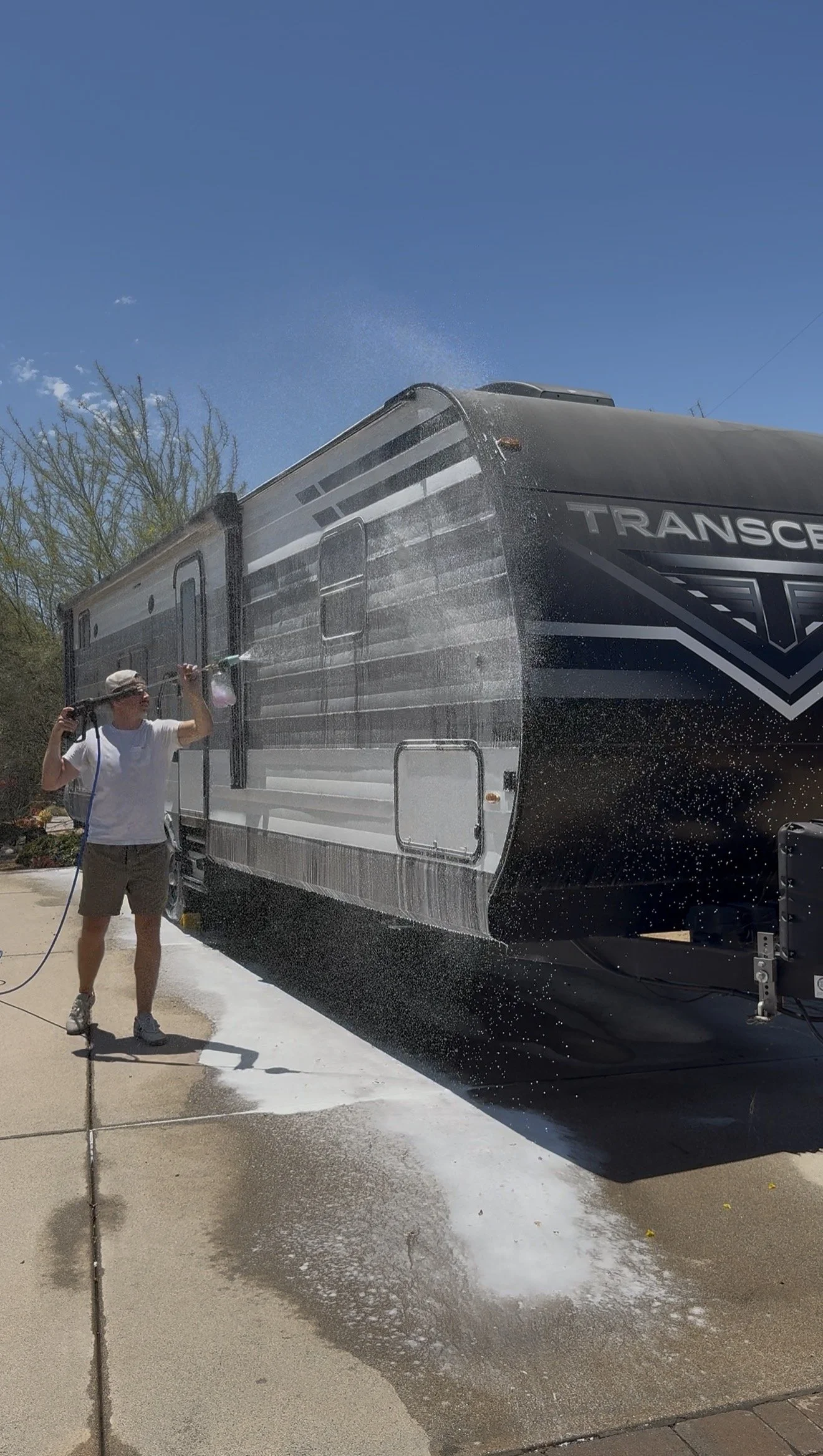A man wearing a white shirt, shorts, and a hat is using a spray wand to wash a large black and gray camper trailer. Water is spraying onto the trailer, creating a mist and foam on the ground below. The trailer has the word 'Transcend' on the side and is parked outdoors with trees and a clear blue sky in the background.