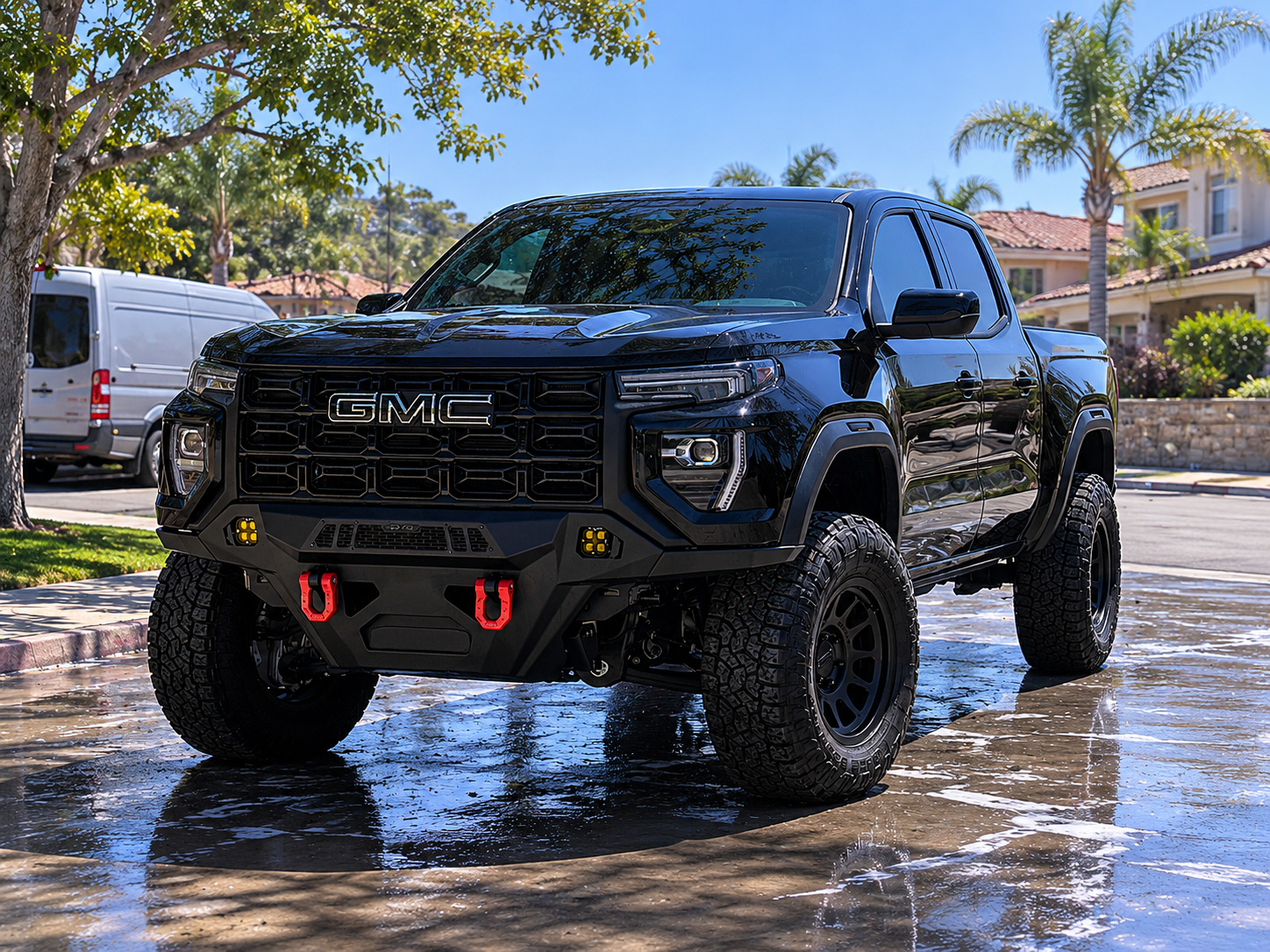 A black GMC off-road pickup truck with large tires, water on the ground, and a residential neighborhood with palm trees in the background.