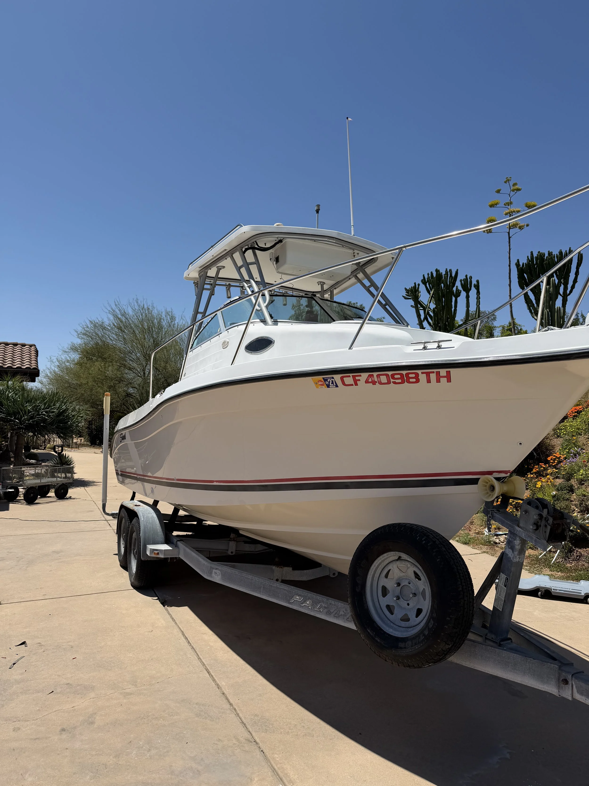 A white boat on a trailer, parked on a concrete driveway with a cactus and desert plants in the background, under a clear blue sky.