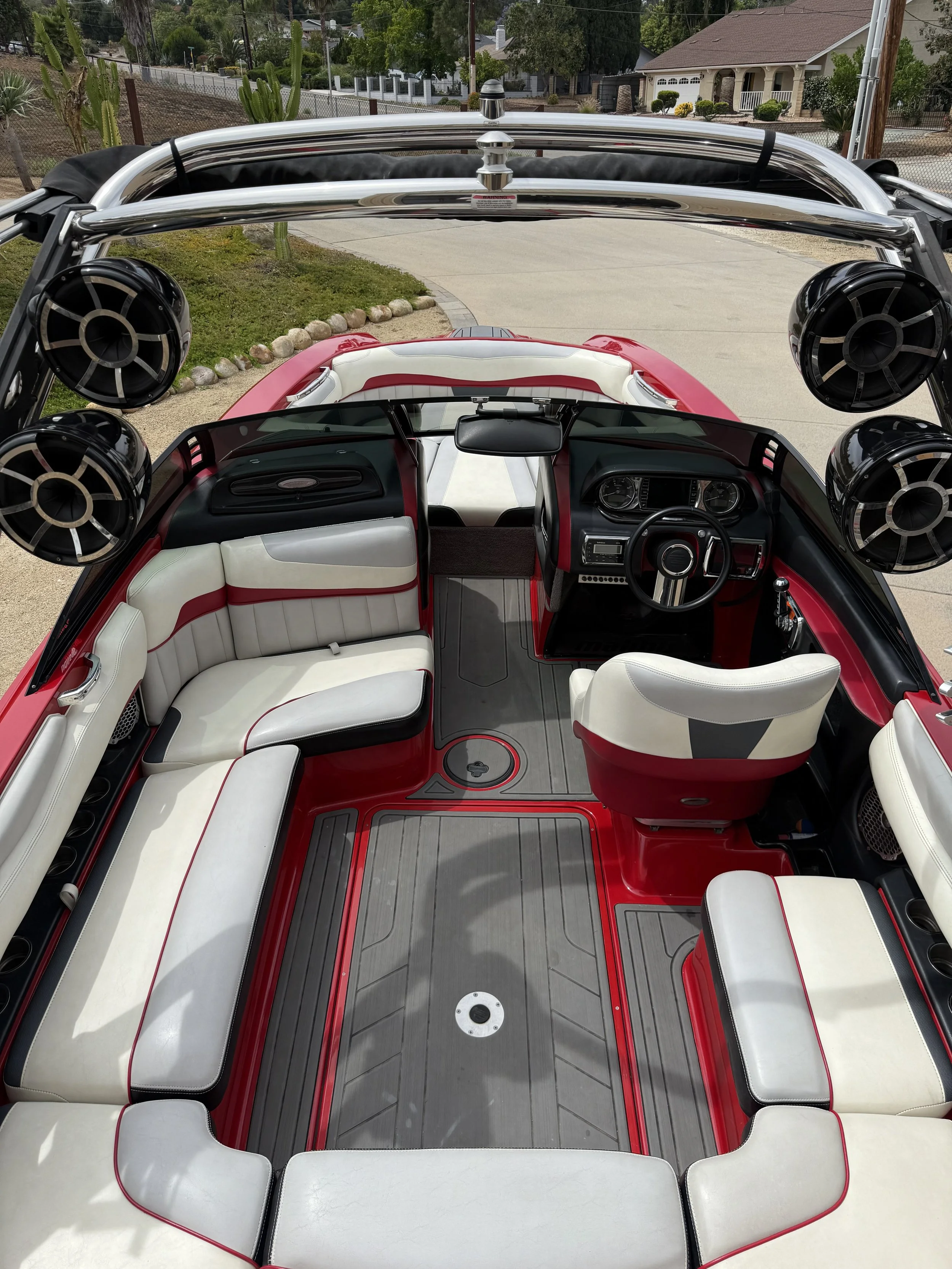 Inside view of a boat with white and red seating, black speakers, and a steering wheel. Cactus plants and houses are visible in the background.