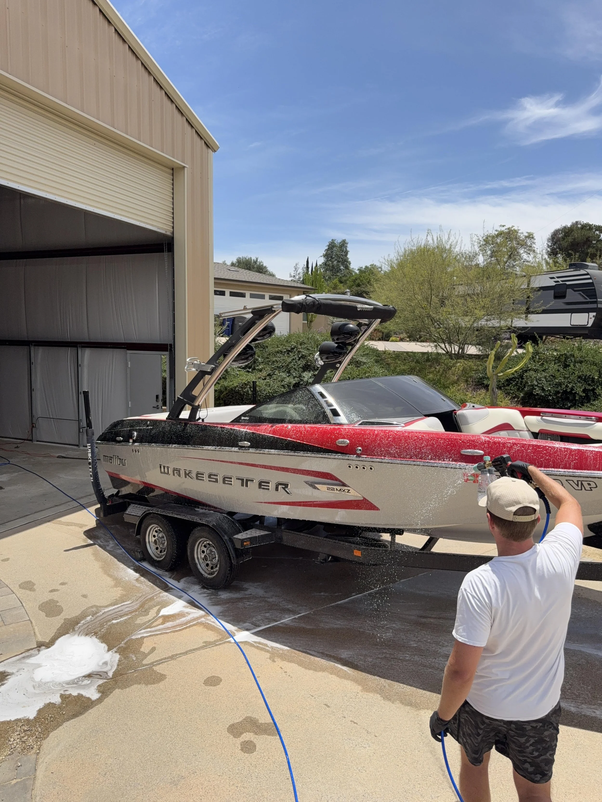 A man in a white t-shirt and camouflage shorts is washing a red and white wakeboard boat on a trailer in a driveway. Soap suds and water are visible on the ground around the vehicle, with a garden hose in use. A partially open garage and a recreational vehicle are visible in the background under a partly cloudy sky.