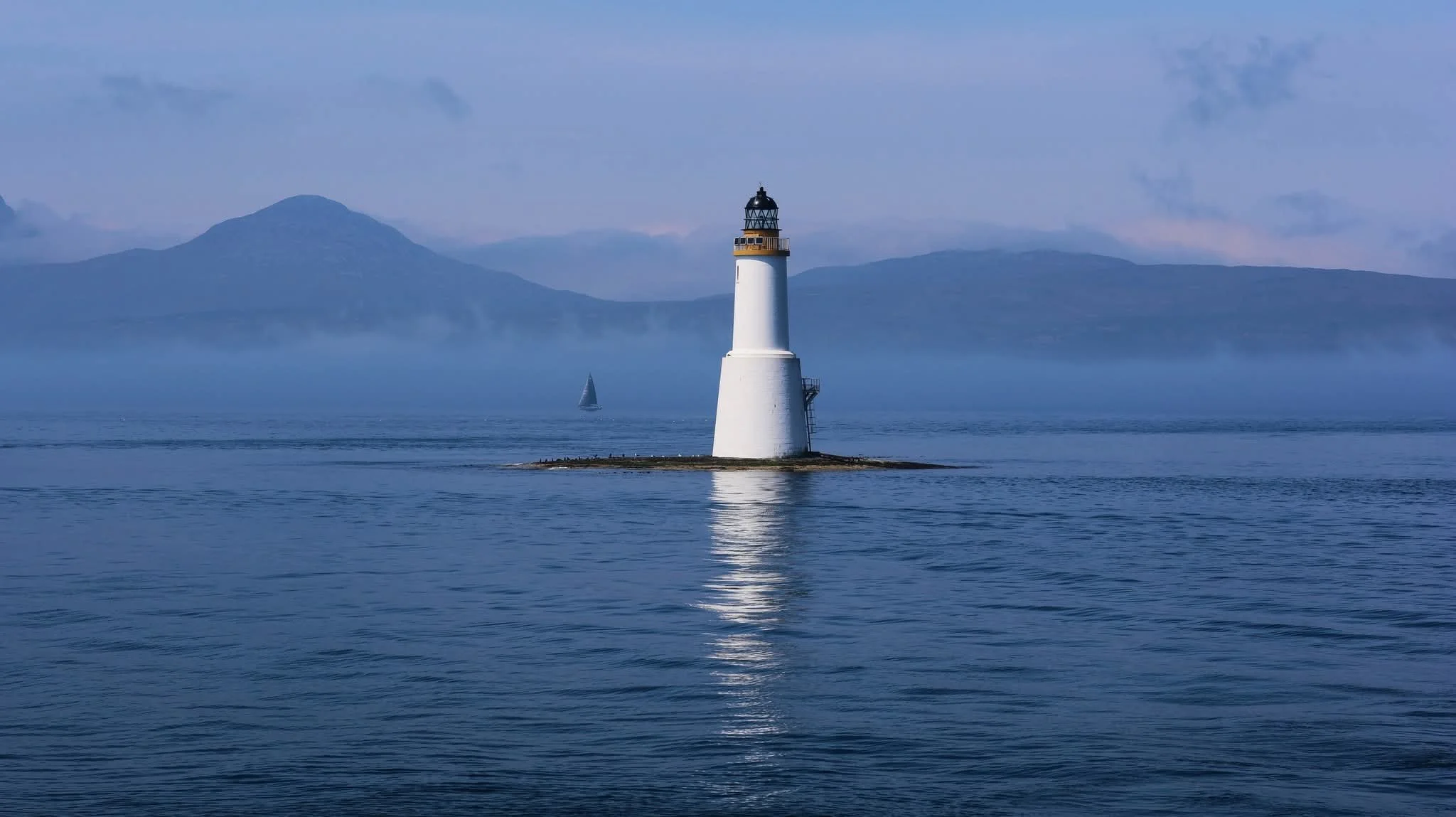 A lighthouse on a small rocky island surrounded by calm water with a sailboat and mountains in the background under a cloudy sky.