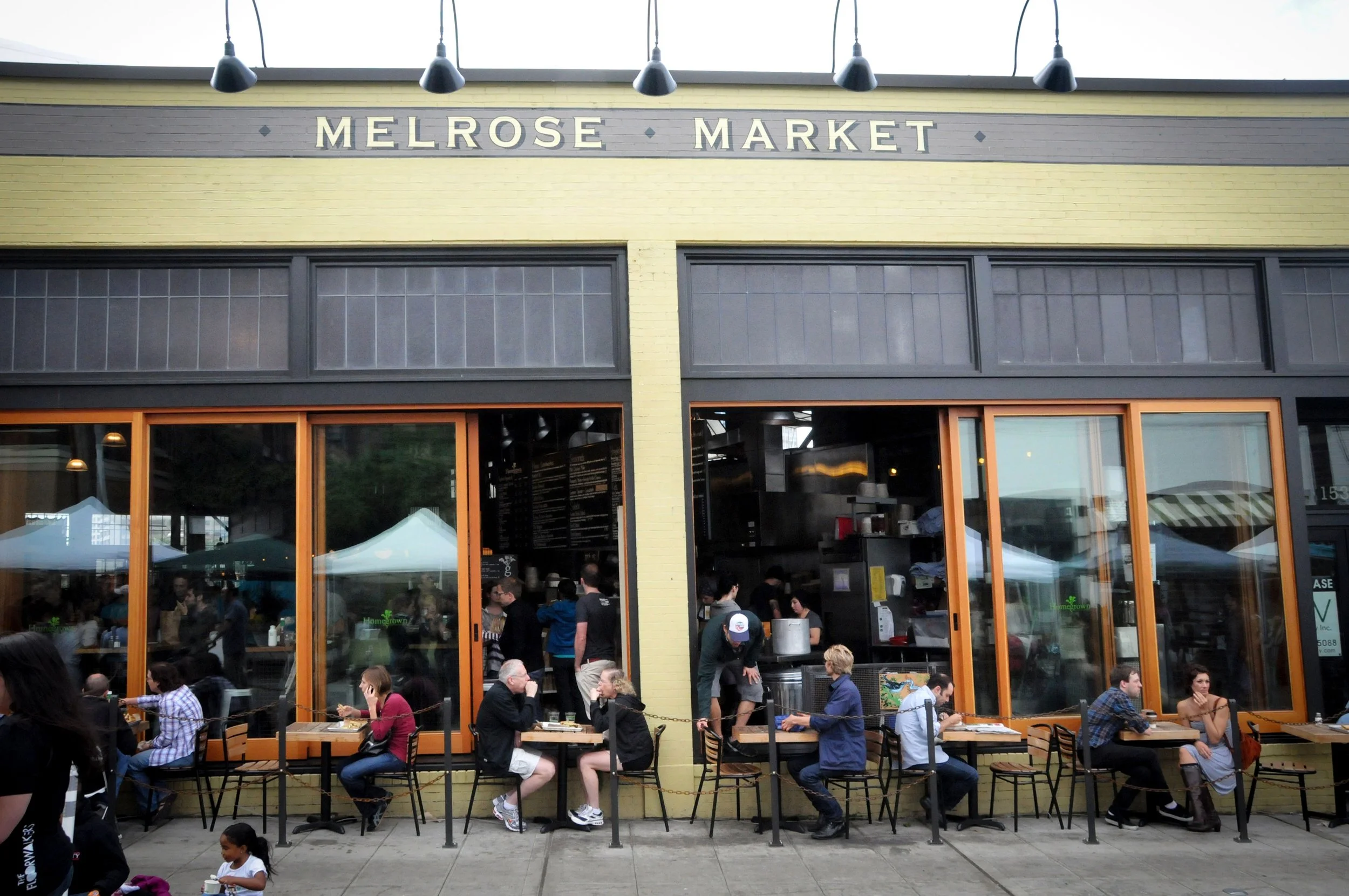 People dining outside Melrose Market, a restaurant with large glass windows and a yellow brick exterior, under hanging lights.