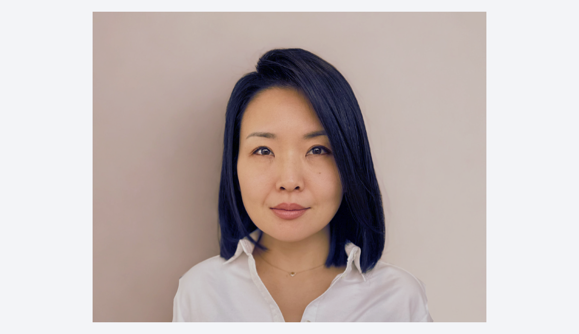 A woman with shoulder-length dark hair and light makeup, wearing a white collared shirt, standing against a plain beige background.