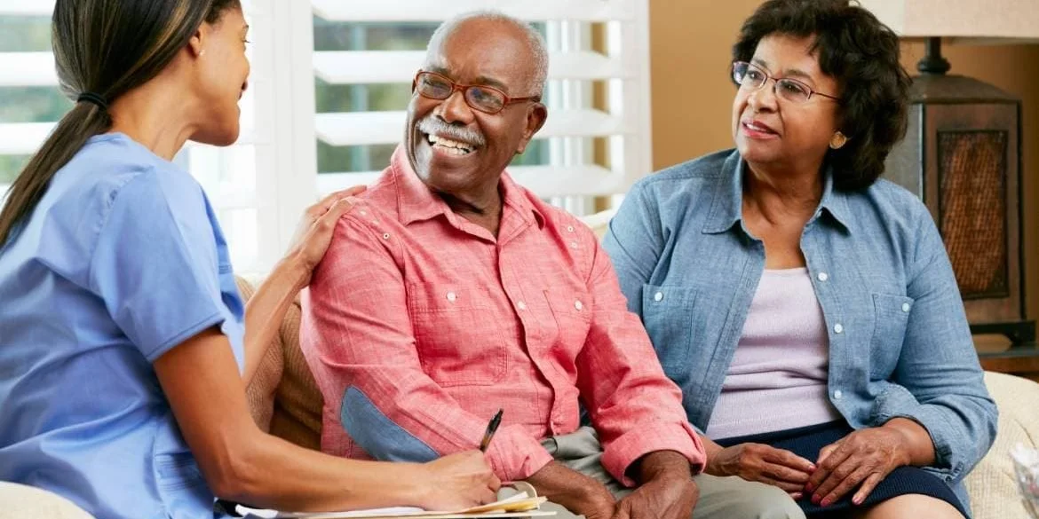 A smiling female nurse talking to an elderly couple on a couch in a living room.