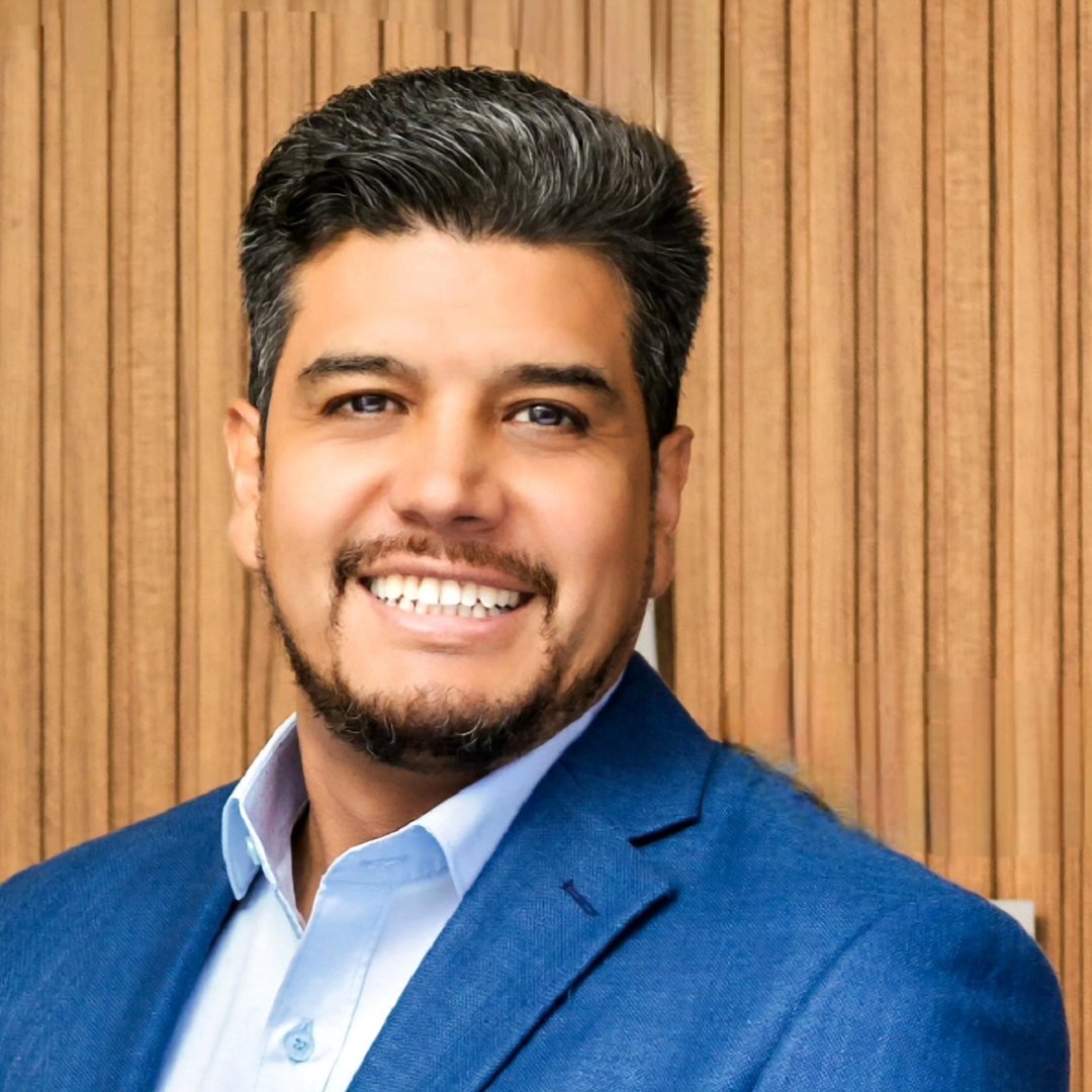 A smiling man with dark hair and a beard, wearing a blue suit jacket and white shirt, sitting in front of a wooden wall.