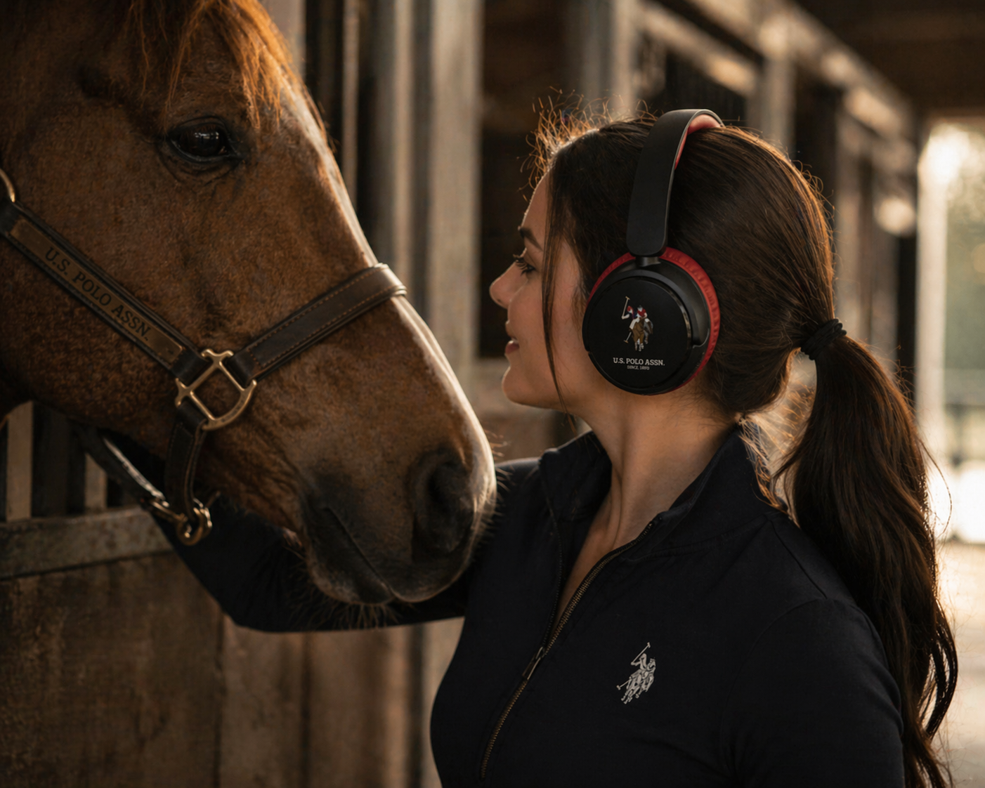 A woman wearing U.S. Polo Assn. headphones looks at a horse in a stable.