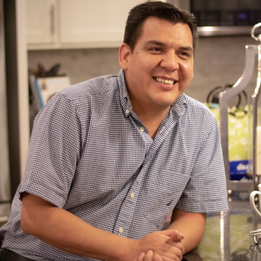 Smiling man in a short-sleeved, button-up shirt in a kitchen with shelves and containers in the background.