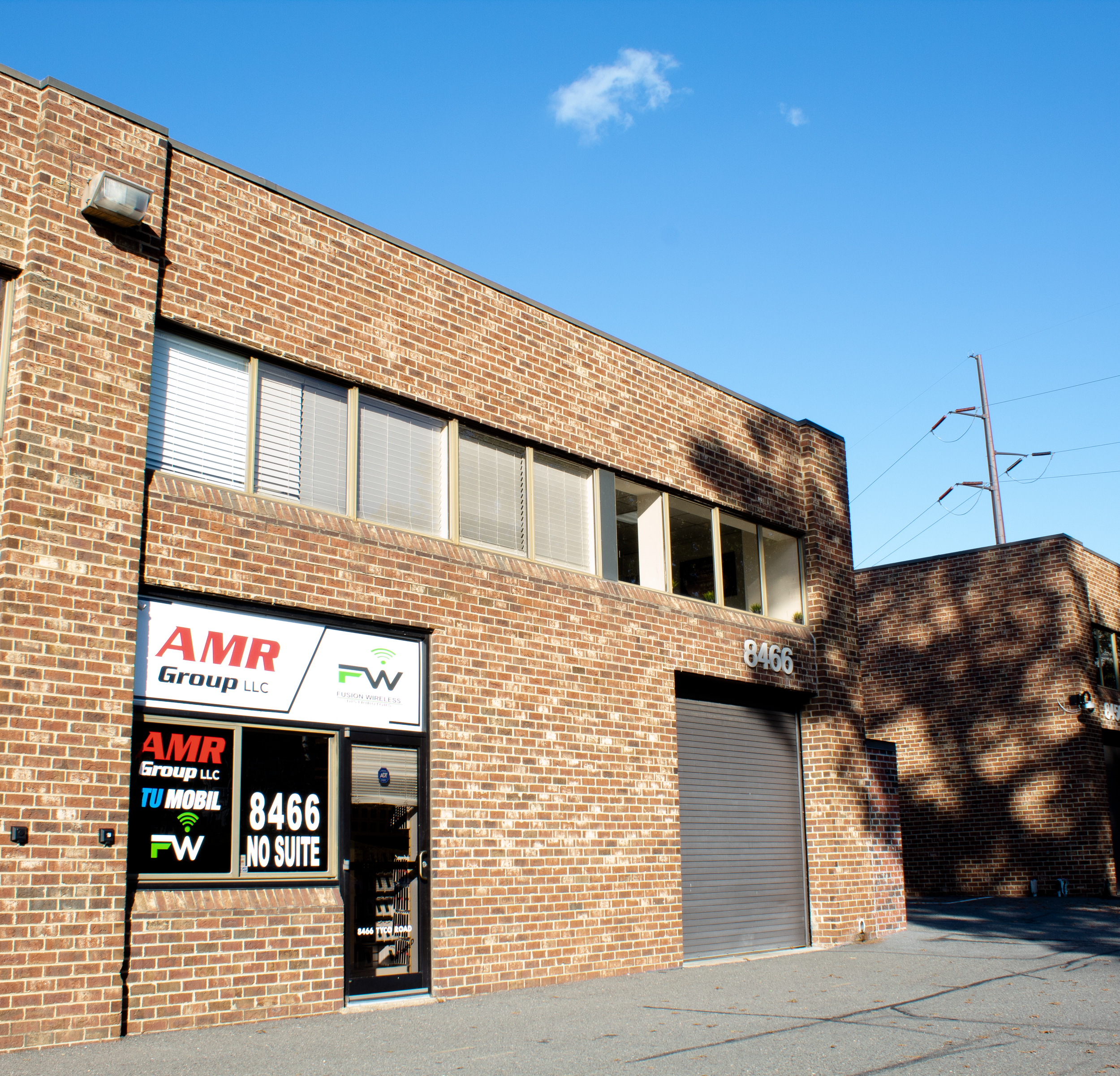 Brick building with a sign displaying various telecommunications and wireless service provider logos, including AMR Group LLC and Fusion Wireless, and large windows, under a clear blue sky with a few small clouds.