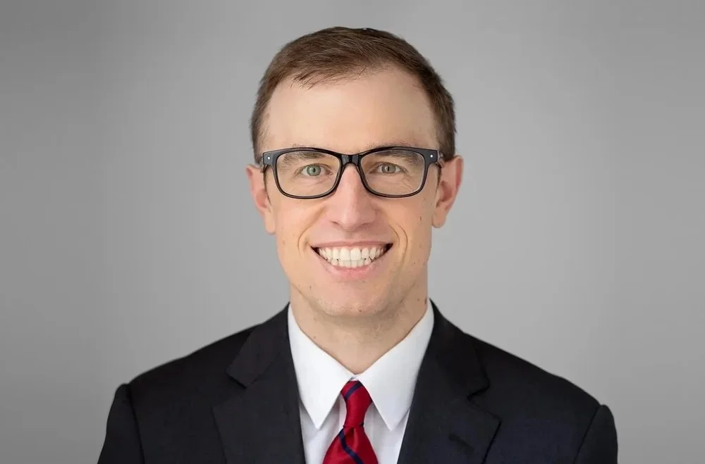 Professional headshot of Zach Remen, Associate Lawyer, Real Estate, wearing smart glasses, black suit, white shirt and red/navy striped tie, smiling.