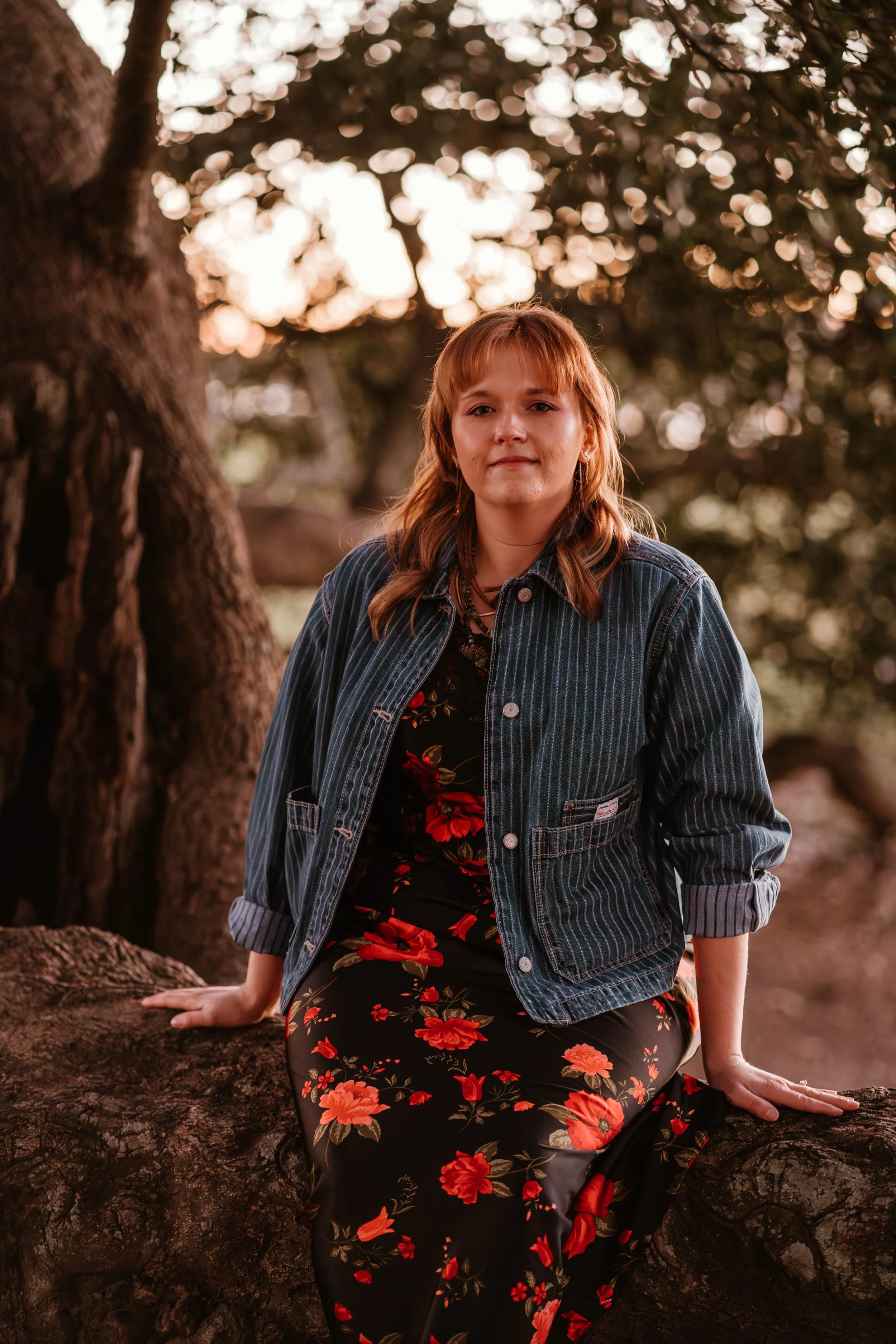 A woman with shoulder-length reddish hair is sitting on a large tree branch outdoors during sunset. She is wearing a denim jacket over a black floral dress with red and orange flowers. The background features blurred trees and warm light filtering through the leaves.