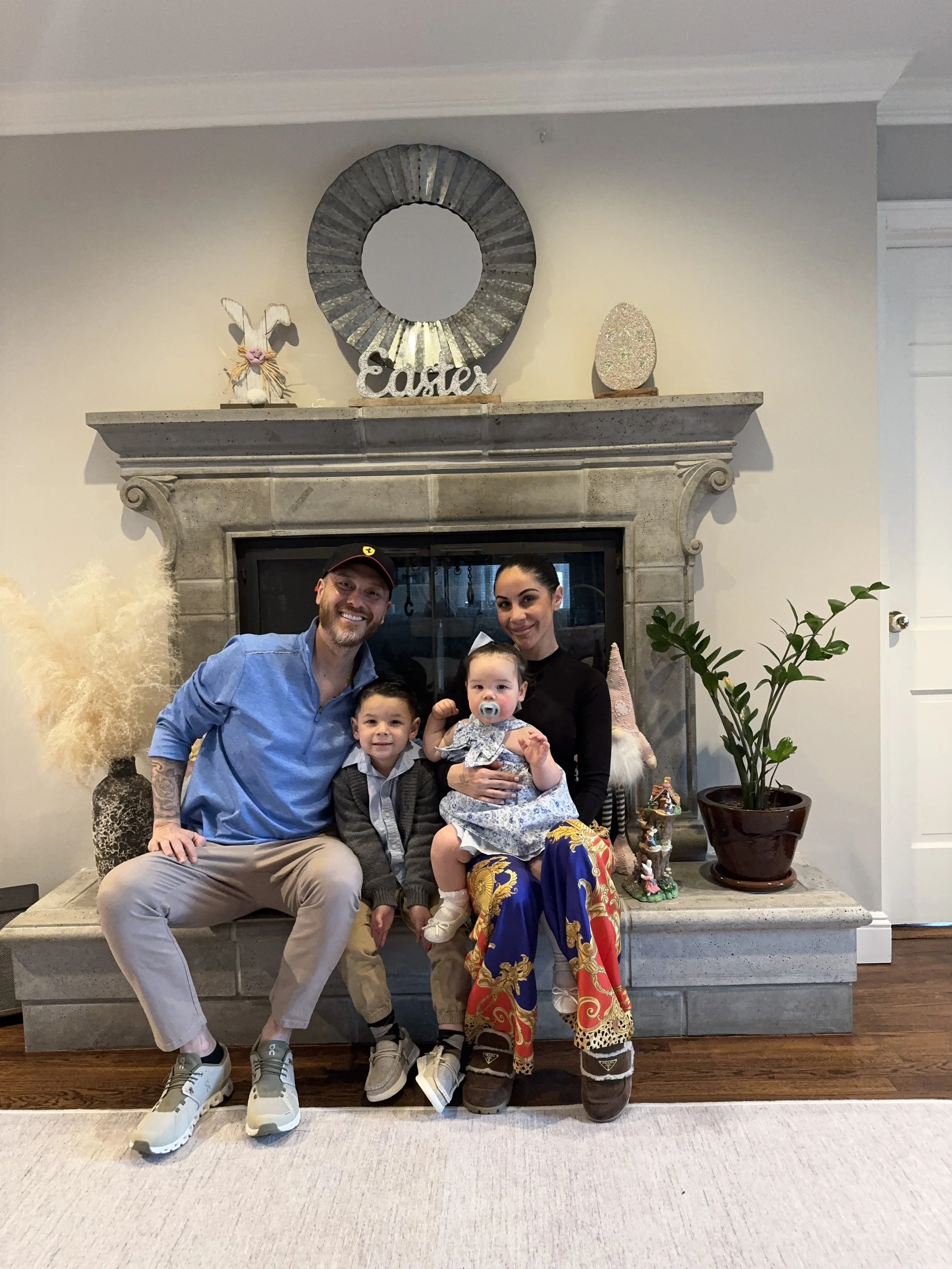 A family of four, including two adults and two children, posing in front of a fireplace decorated with Easter-themed items and a large mirror. The background includes a potted plant and Easter decorations.