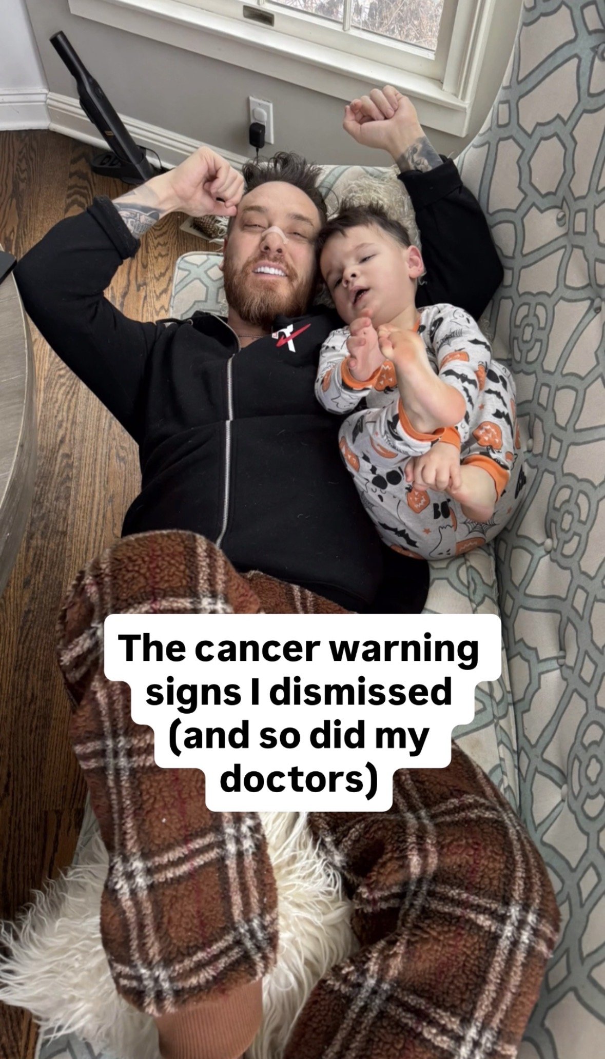 A man and a young boy lying on the floor, smiling with one arm raised, next to a window with blinds. The boy is wearing pajamas with Halloween pumpkins. The man is wearing a black hoodie with a red and white design. Text at the bottom reads, "The cancer warning signs I dismissed (and so did my doctors)."