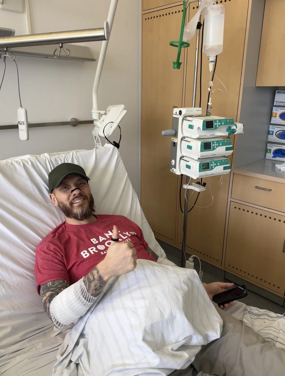 A man with a beard, wearing a baseball cap and a red t-shirt, lying in a hospital bed, smiling and giving a thumbs up. Medical equipment and an IV are visible beside him.