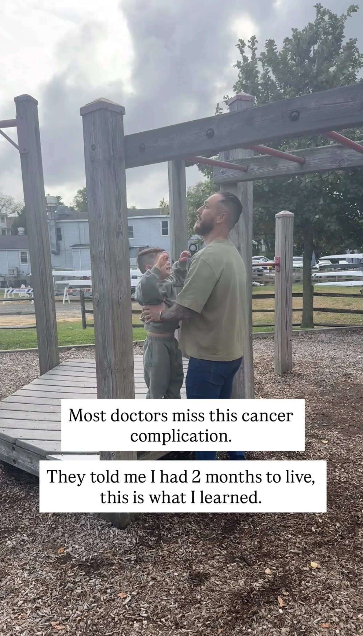 A man lifting a young boy on a playground. The man has a beard and is wearing a beige t-shirt, and the boy is in a gray hoodie and pants. They are standing on a wooden platform near gym equipment, with a playground structure and trees in the background. There are clouds in the sky.