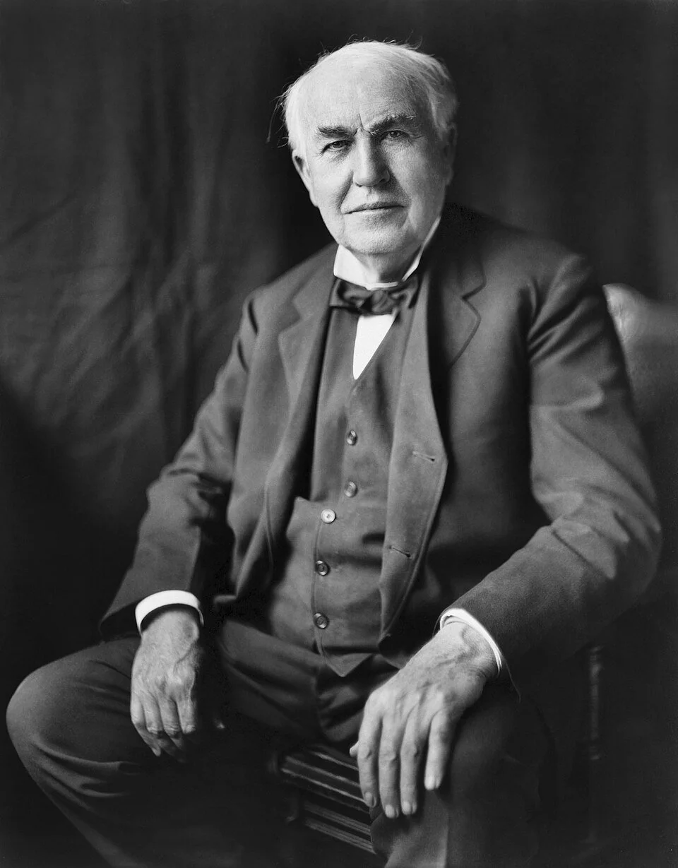 Black and white portrait of an elderly man in a suit with a bow tie, sitting on a wooden chair, looking directly at the camera with a serious expression.