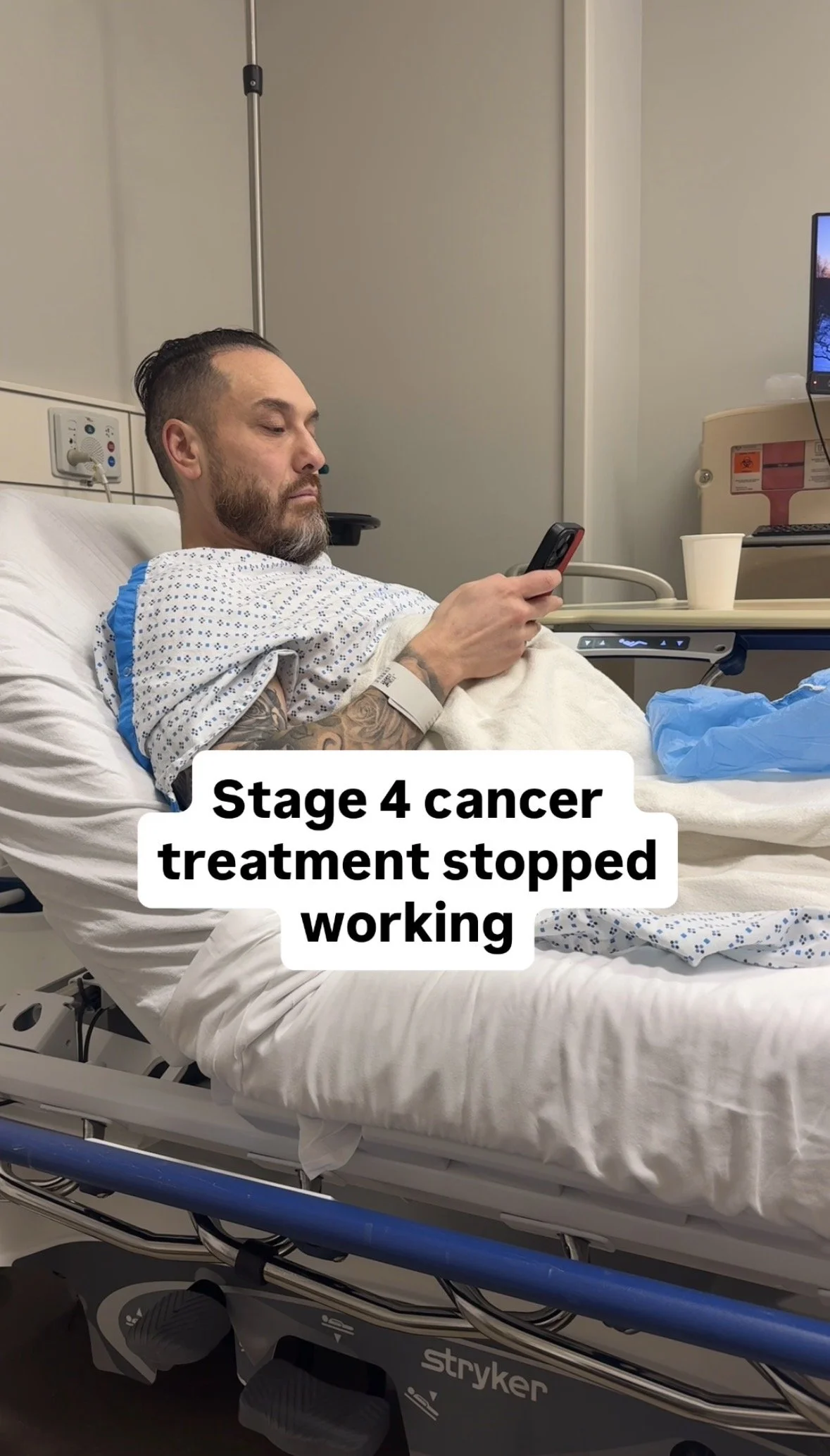 A man with a beard and tattooed arm in a hospital bed, wearing a hospital gown, looking at his phone. The hospital room has medical equipment, a monitor, and a cup on a tray. Text overlay reads, "Stage 4 cancer treatment stopped working."