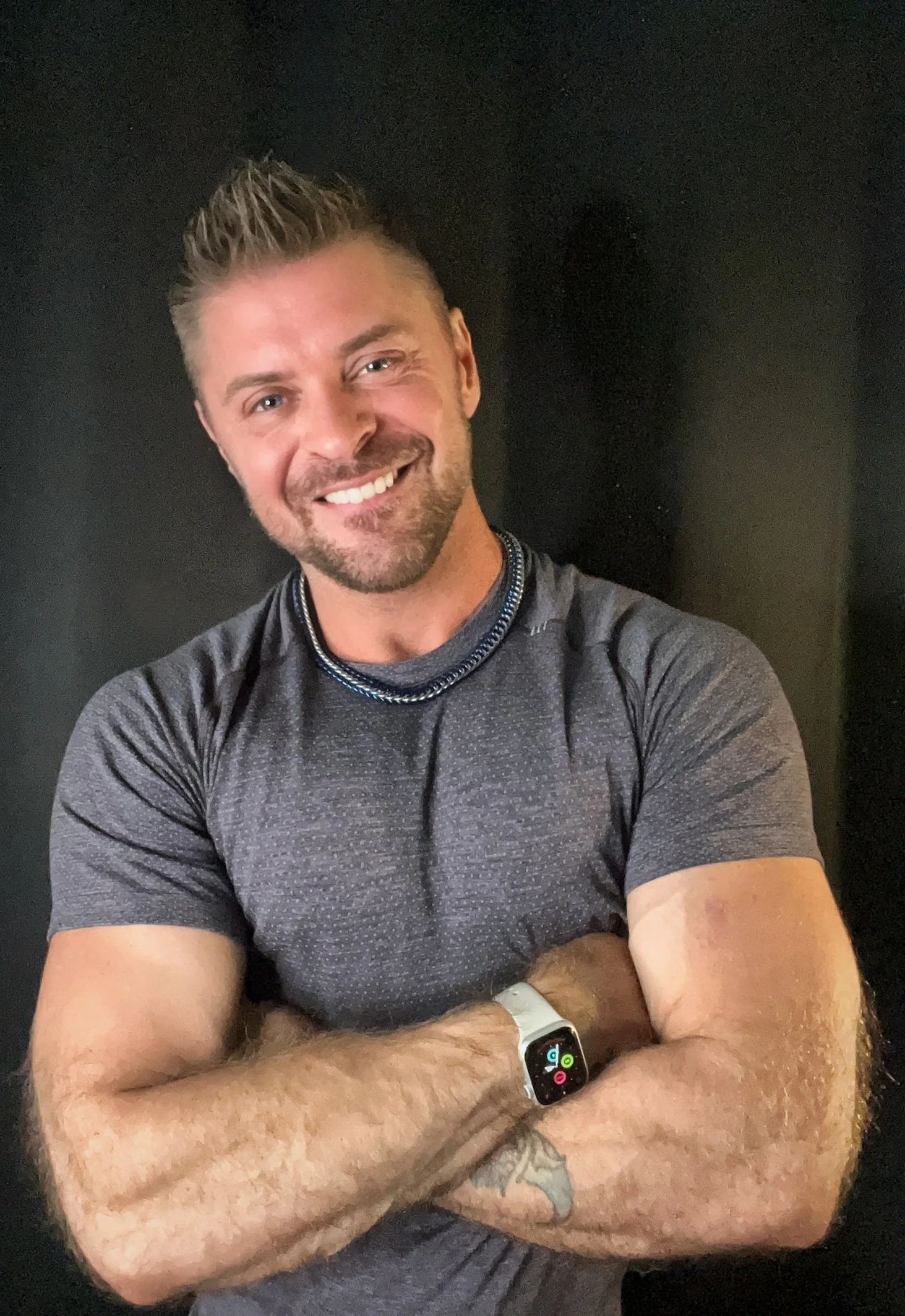 A smiling man with light skin, short spiked hair, and a beard, wearing a gray athletic shirt, a smartwatch, and a necklace, sitting with crossed arms against a dark background.
