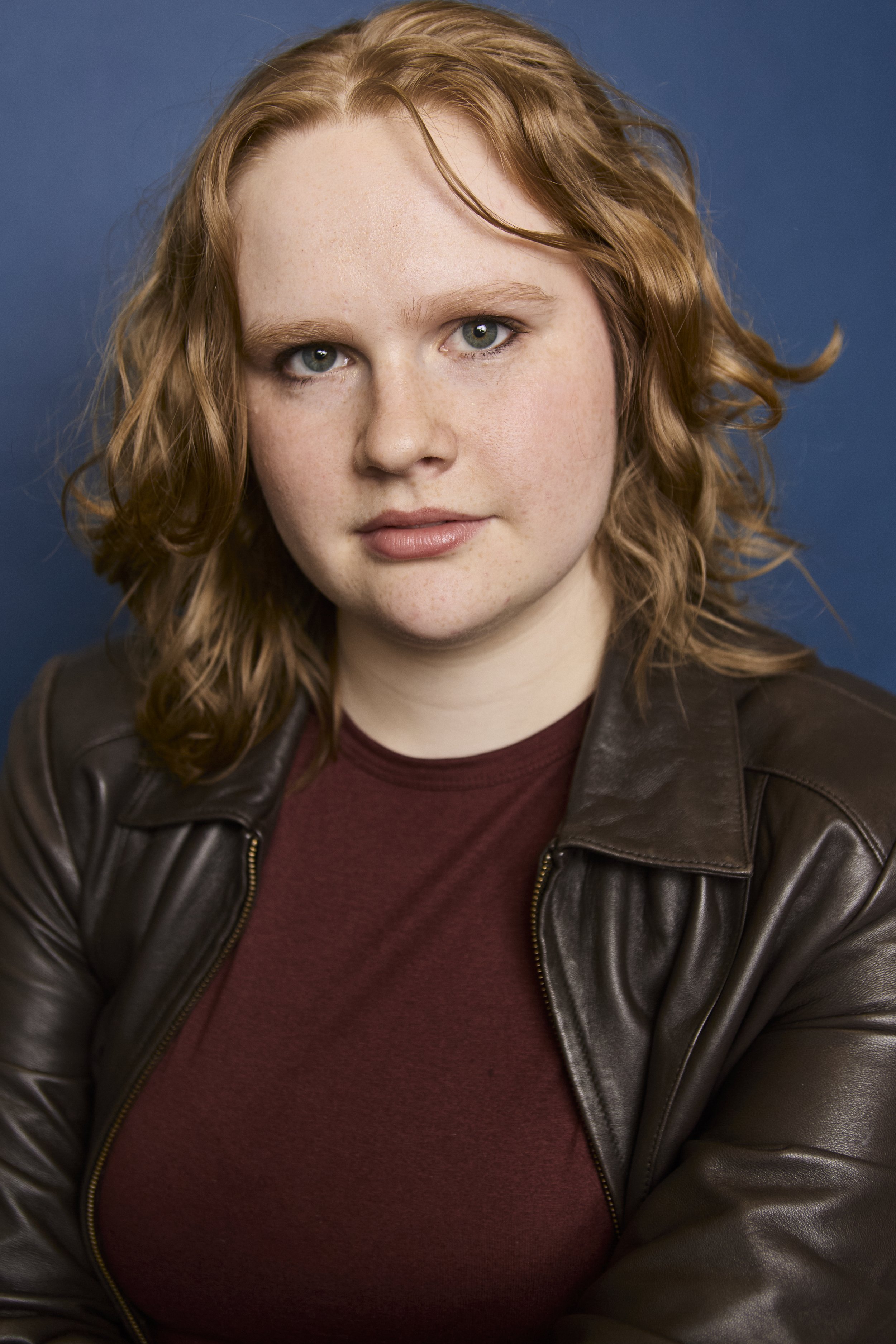 A close-up portrait of a young woman with red hair, blue eyes, and fair skin, wearing a dark brown leather jacket over a maroon shirt, against a solid blue background.