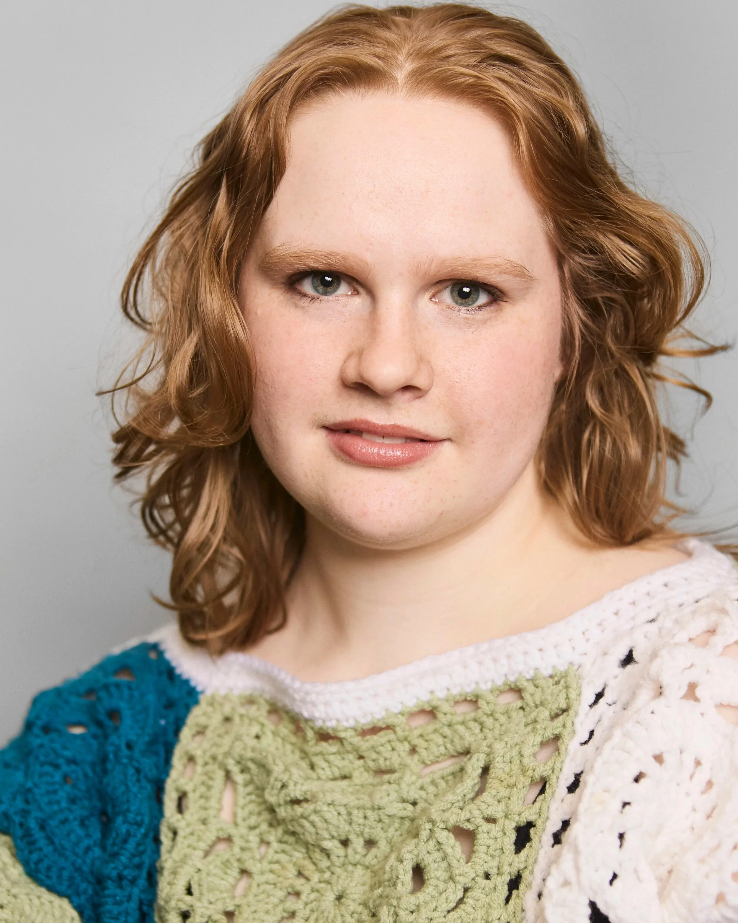 A close-up portrait of a woman with red curly hair, blue eyes, and light skin, smiling softly, wearing a multicolored crocheted sweater.