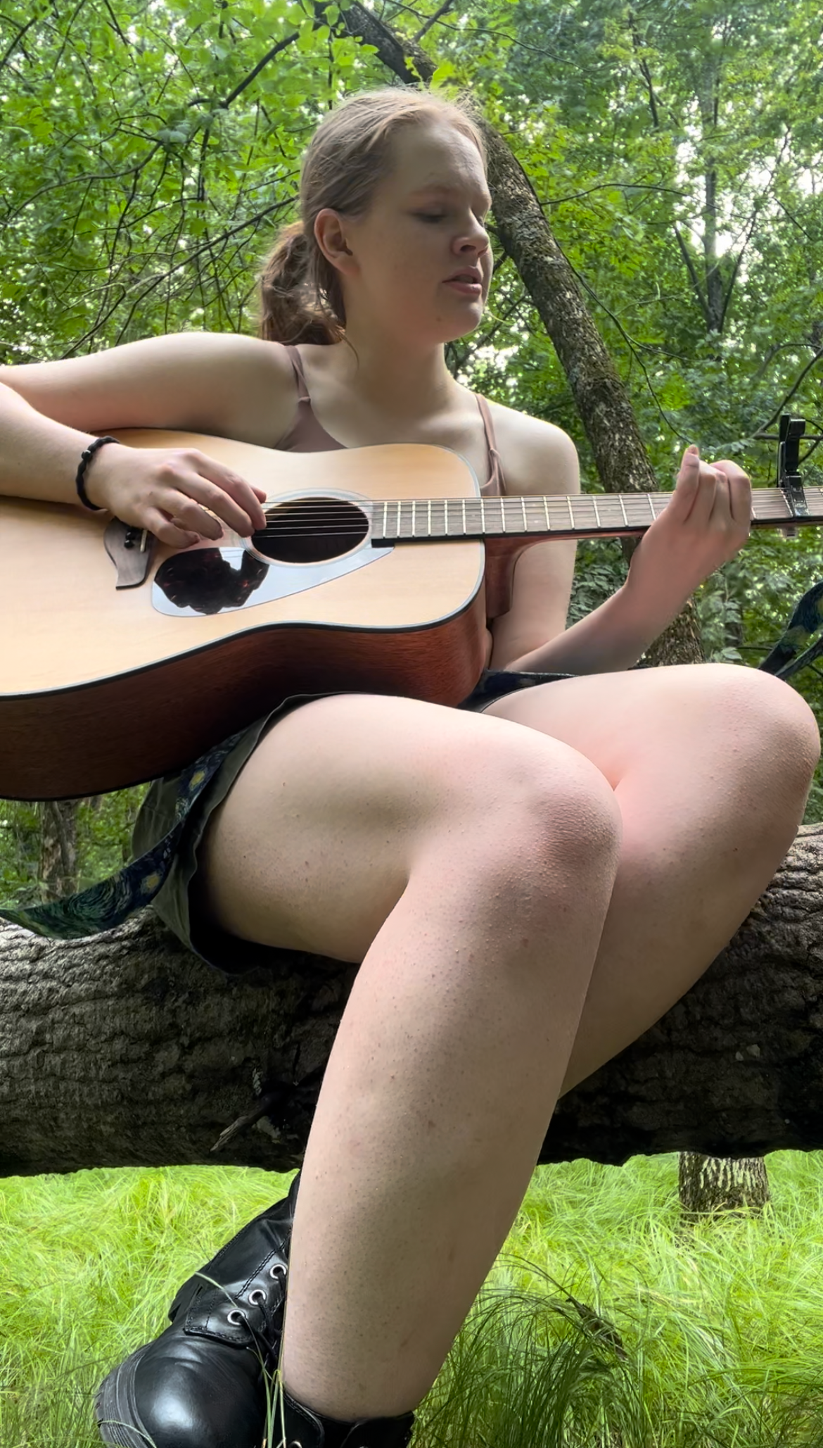 A young woman sitting on a tree branch in a forest, playing an acoustic guitar, with her legs and boots visible.