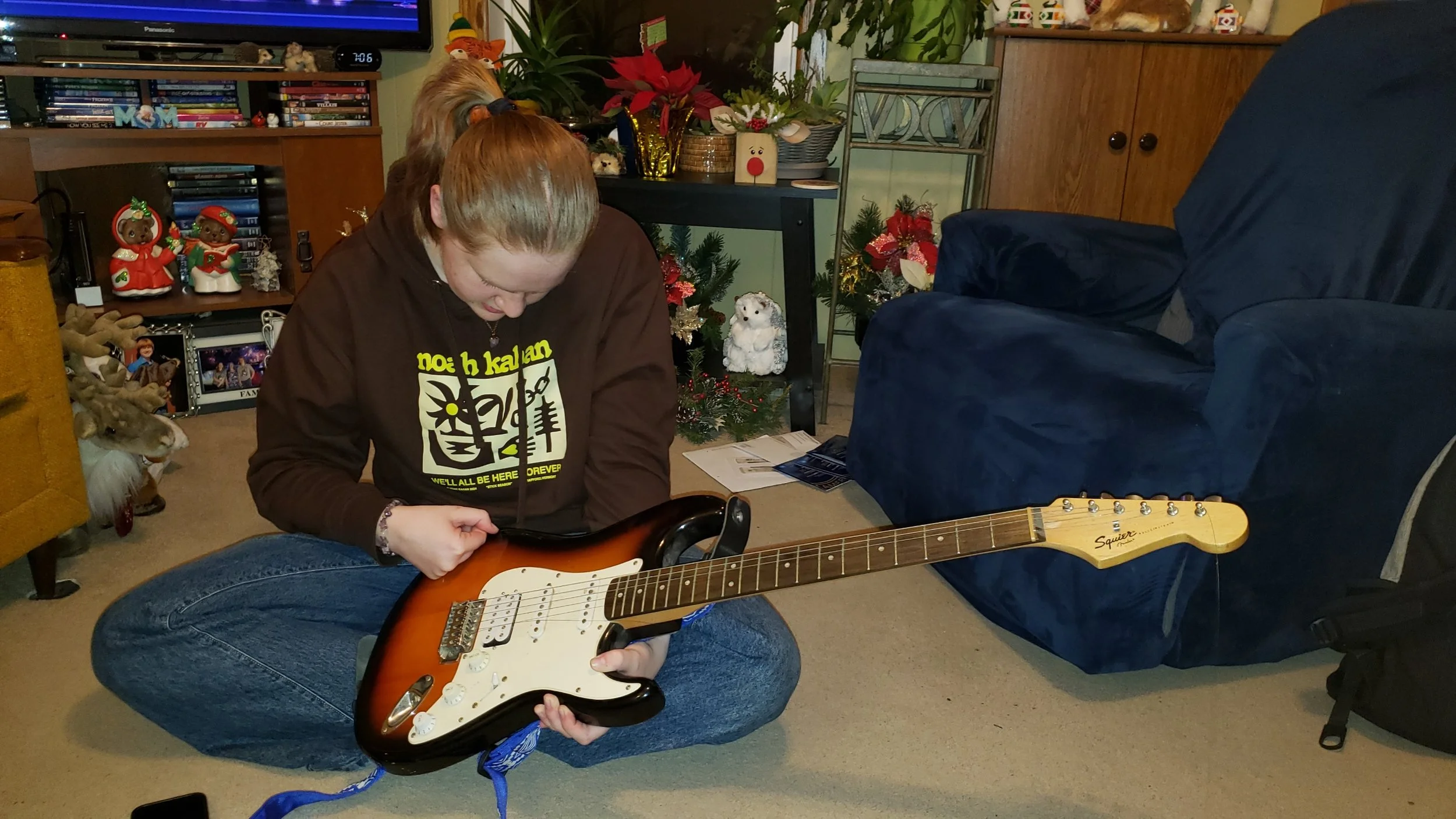 Person sitting on the floor playing an electric guitar in a living room decorated for Christmas, with Christmas trees, ornaments, and holiday decorations around.