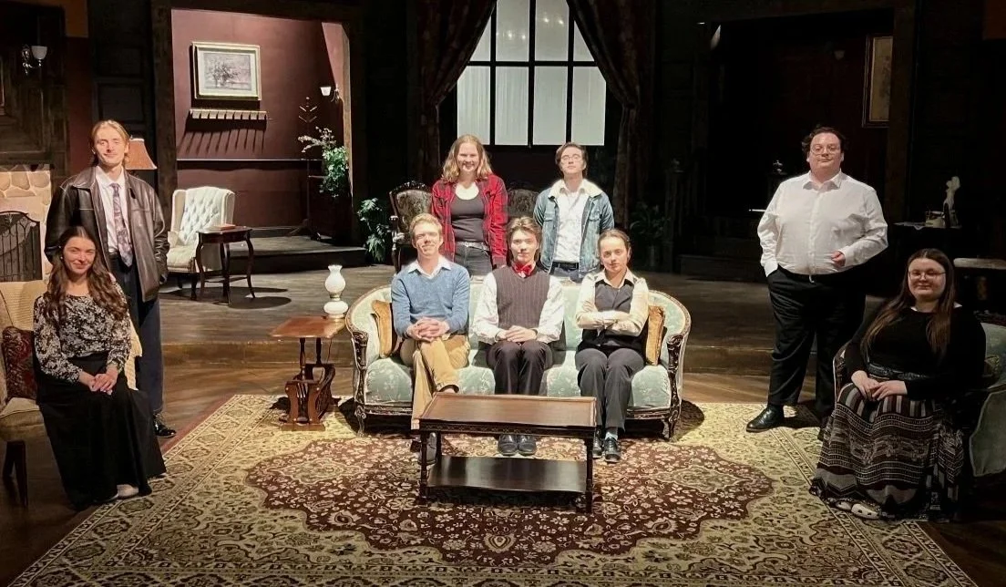 Group of nine people gathered in a living room, some sitting on a sofa and others standing around, with mid-century style furniture and dark wood paneling.