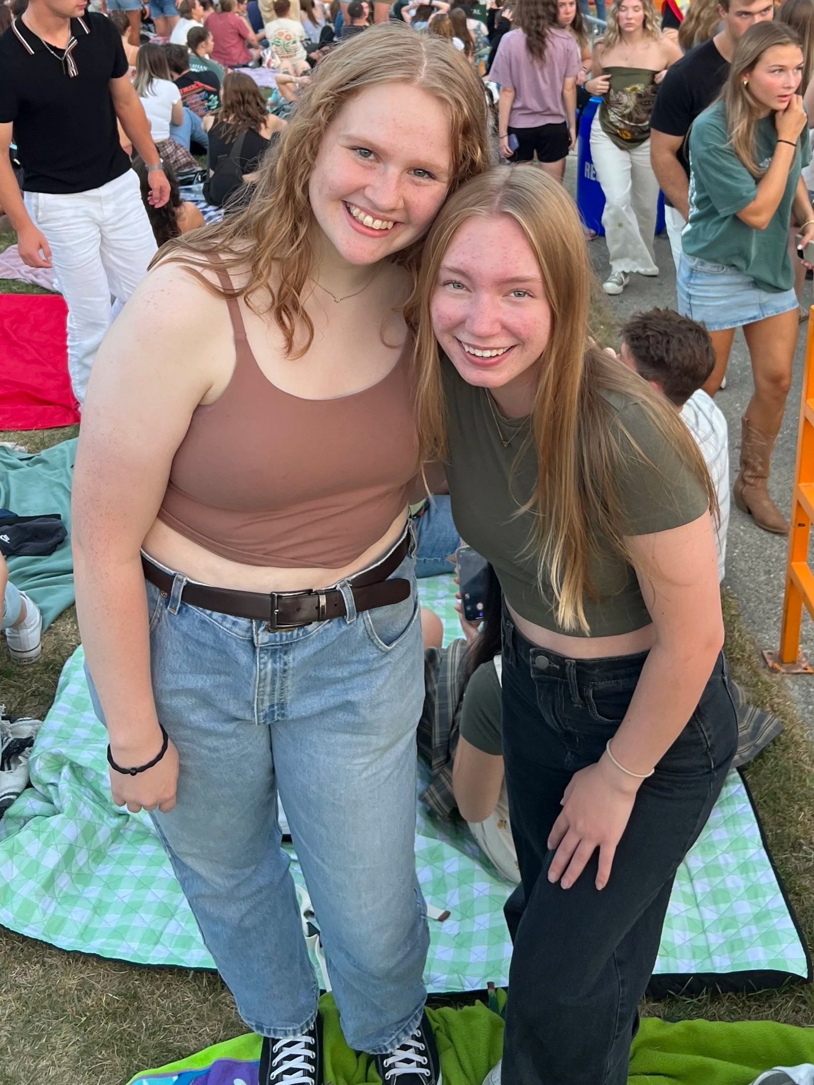 Two young women with red hair smiling and posing together at an outdoor event with many people in the background.