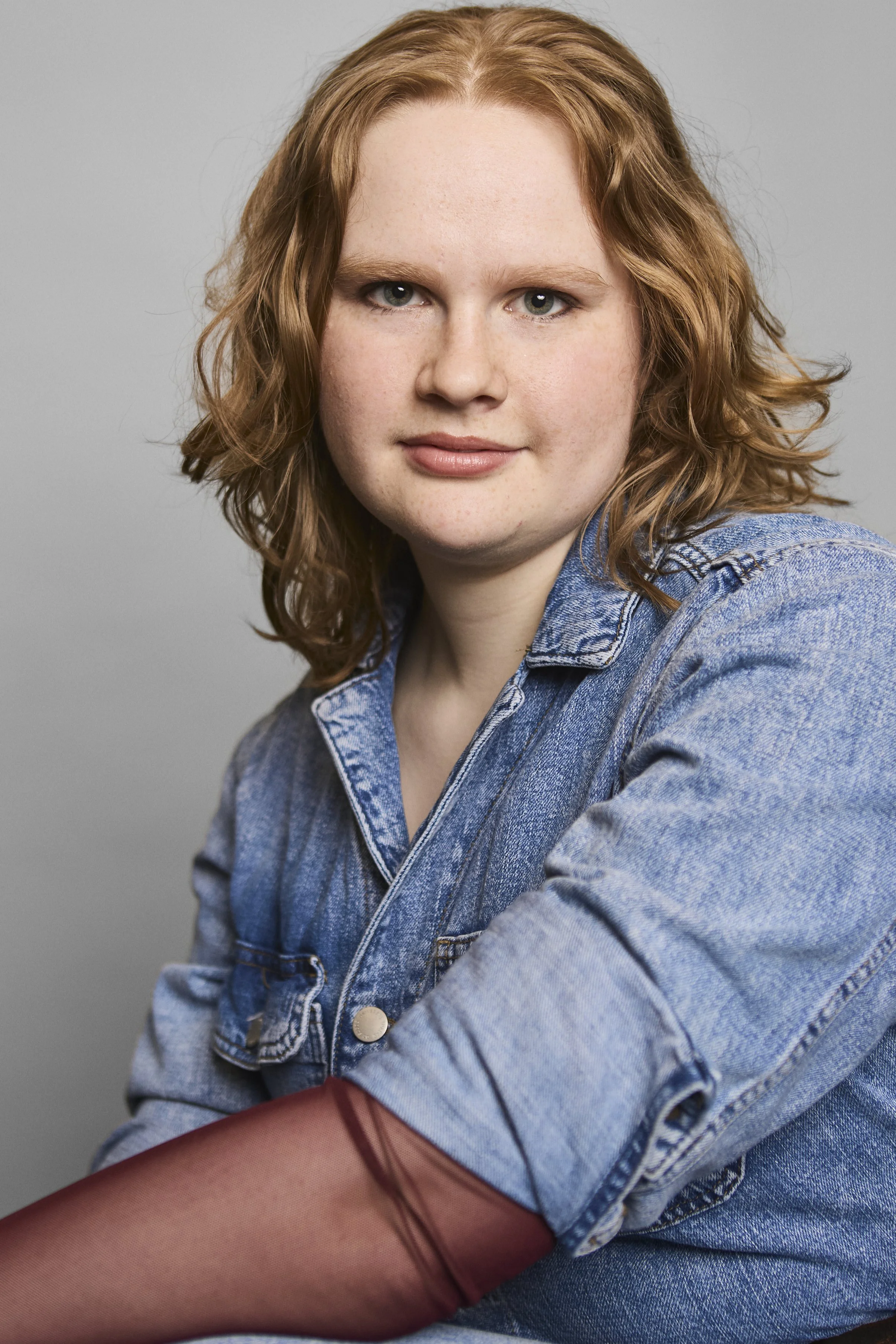 A young woman with shoulder-length wavy red hair and blue eyes wearing a denim jacket over a red top, sitting against a gray background.