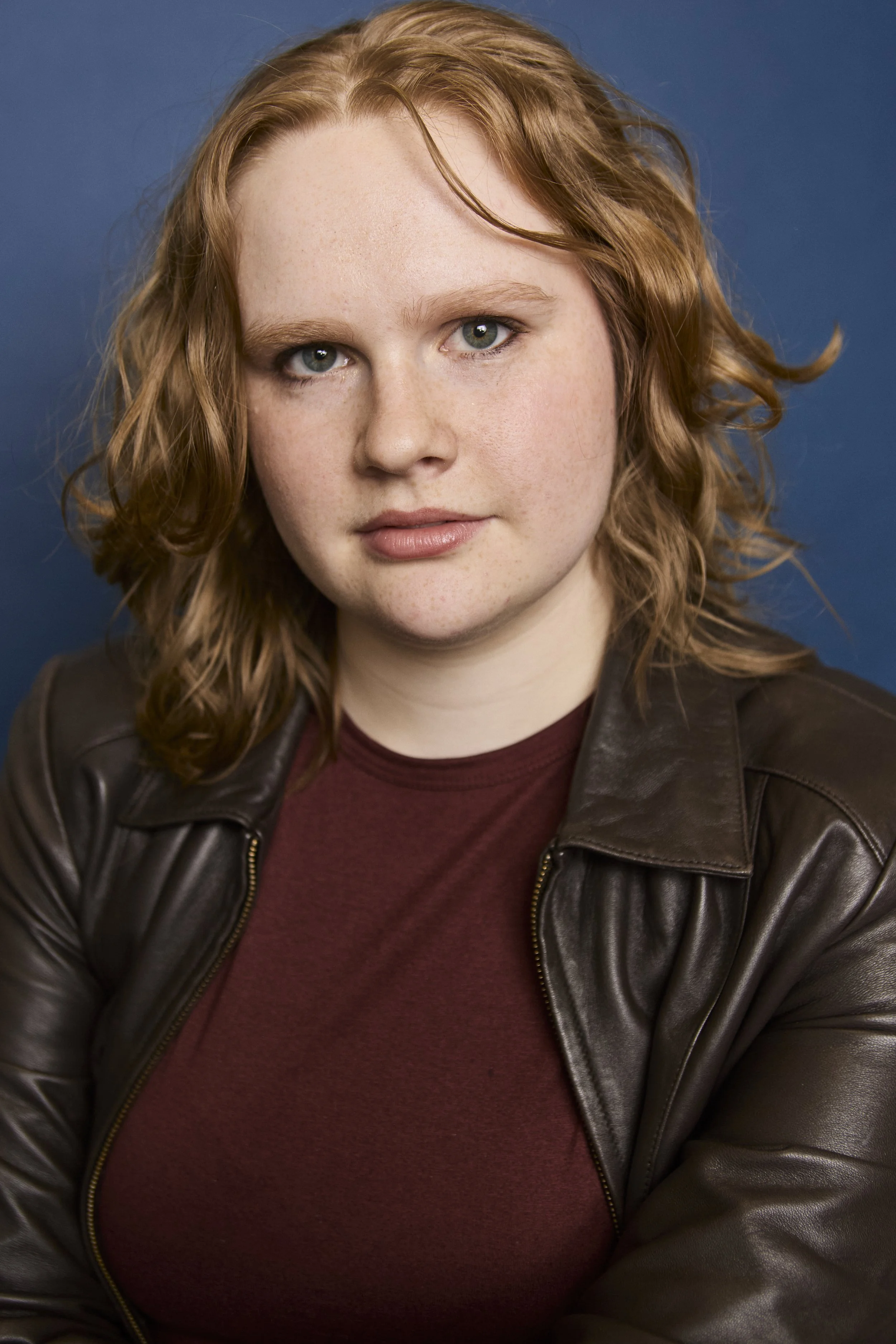 Close-up of a woman with wavy red hair, light skin, and blue eyes wearing a dark leather jacket over a maroon shirt, against a blue background.
