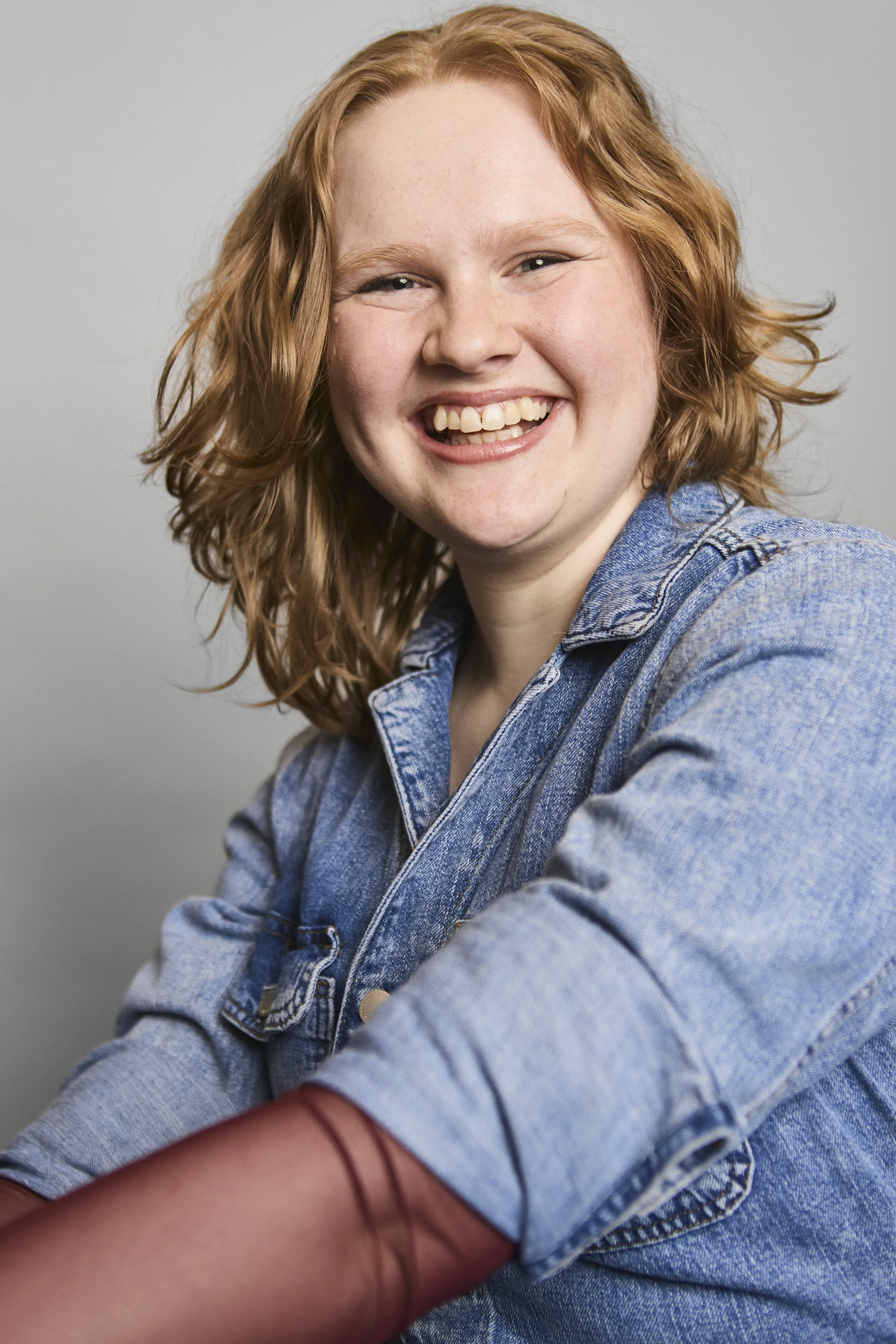 A young woman with curly red hair and a wide smile wearing a denim jacket, leaning forward and resting her arm on a surface.