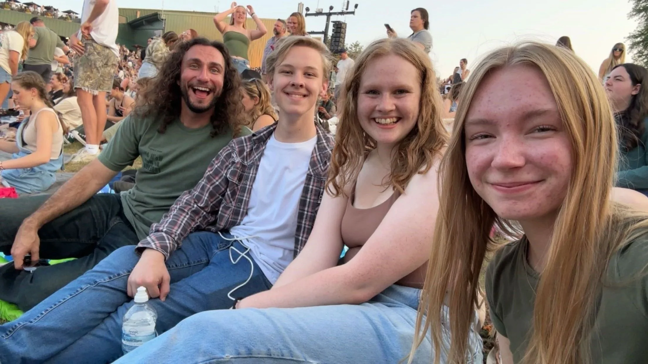 A group of four young people, three girls and one boy, sitting outdoors at a crowded event, smiling at the camera. Other people are gathered in the background, some standing and some seated.