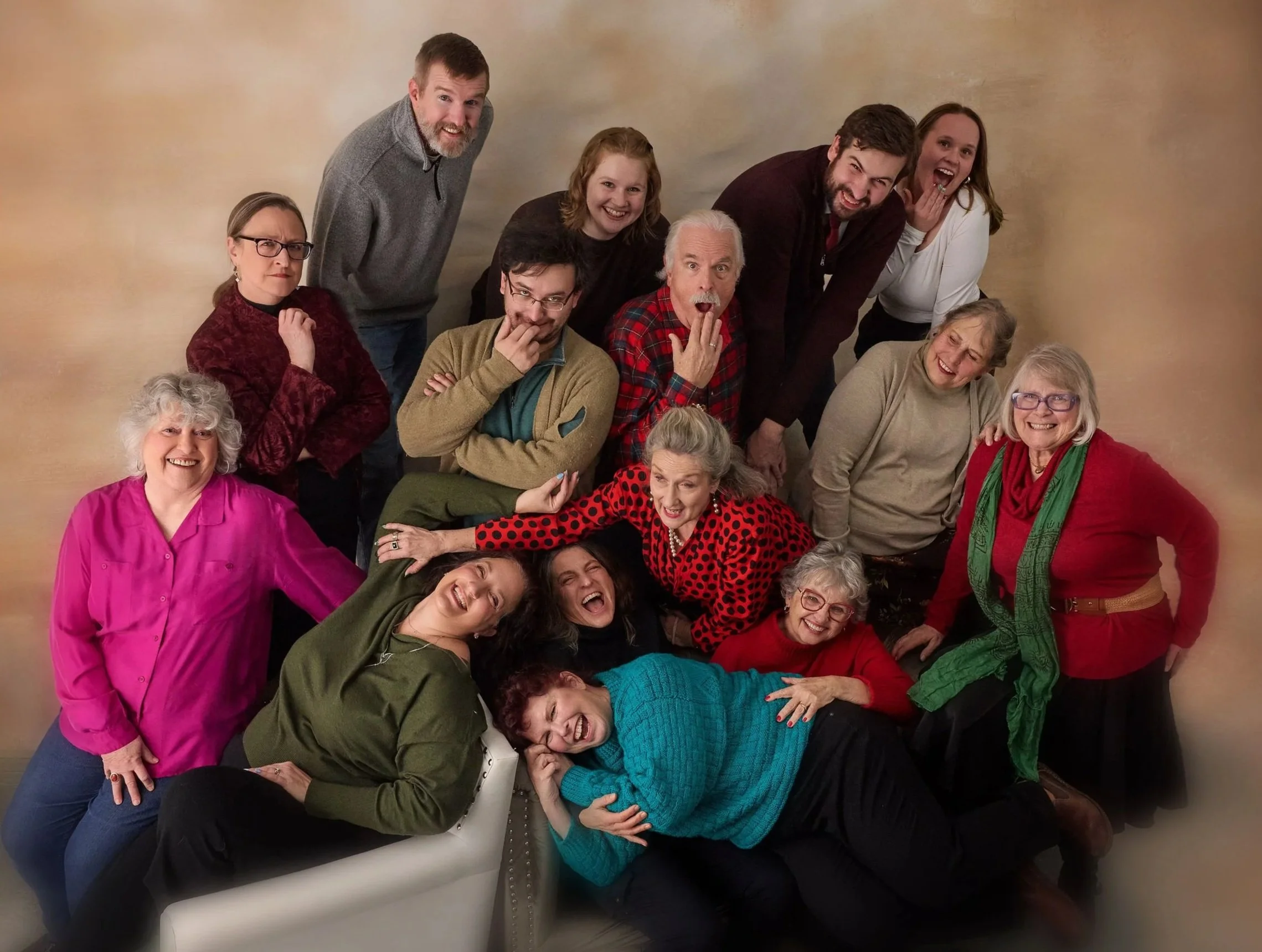 A large group of twelve people, of various ages, are posing playfully together in a casual setting, some standing, some sitting or leaning, against a light-colored background. They are smiling and making fun expressions.