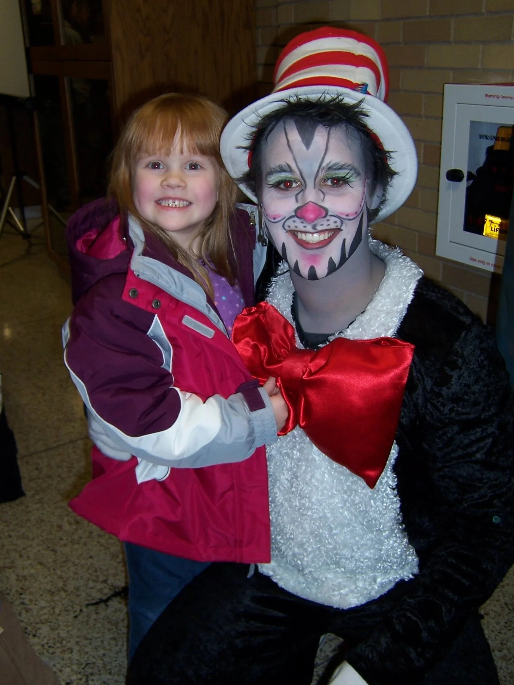 A young girl with red hair, smiling, standing next to a person dressed as the Cat in the Hat with makeup and costume, in an indoor setting.