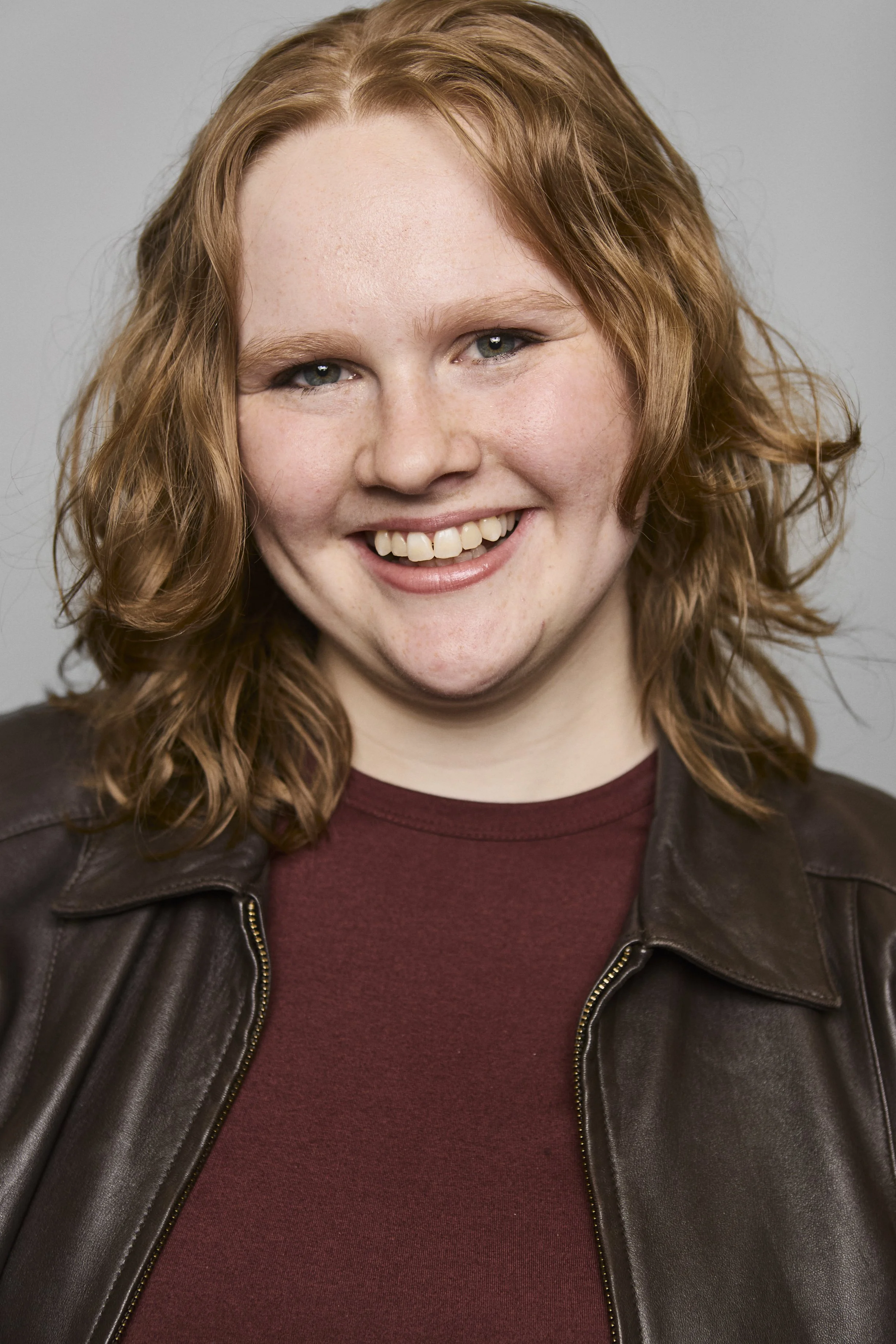 A young woman with red curly hair smiling, wearing a maroon shirt and a brown leather jacket.
