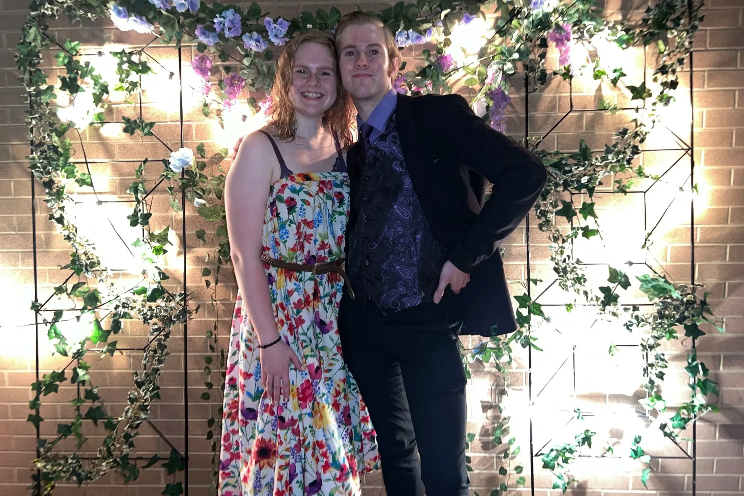 A smiling young woman with reddish curly hair in a colorful floral dress standing next to a young man with reddish hair in a dark suit jacket and patterned shirt, both posing in front of a decorative wall with lights, flowers, and bushy greenery.