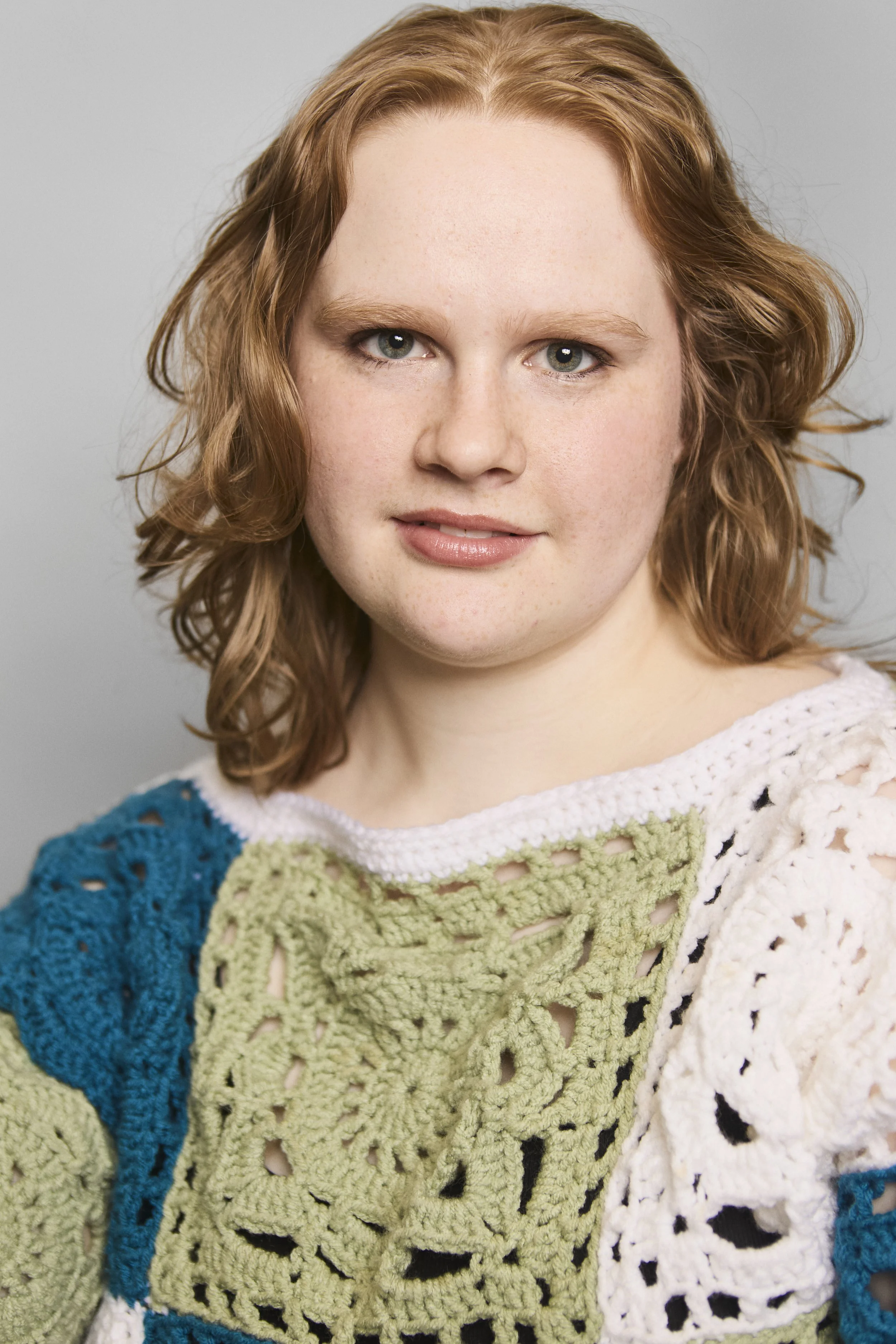 A close-up portrait of a woman with red hair, blue eyes, and freckles wearing a multicolored, crocheted sweater with green, white, blue, and black patterns.
