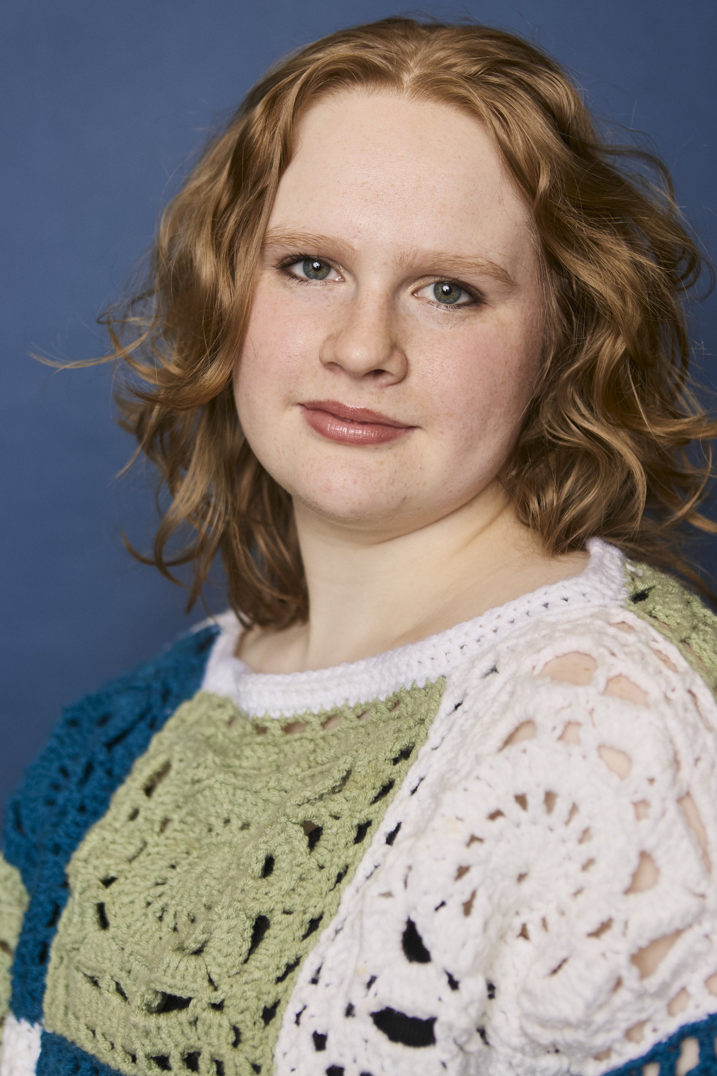 Close-up of a woman with curly red hair looking at the camera against a plain blue background.