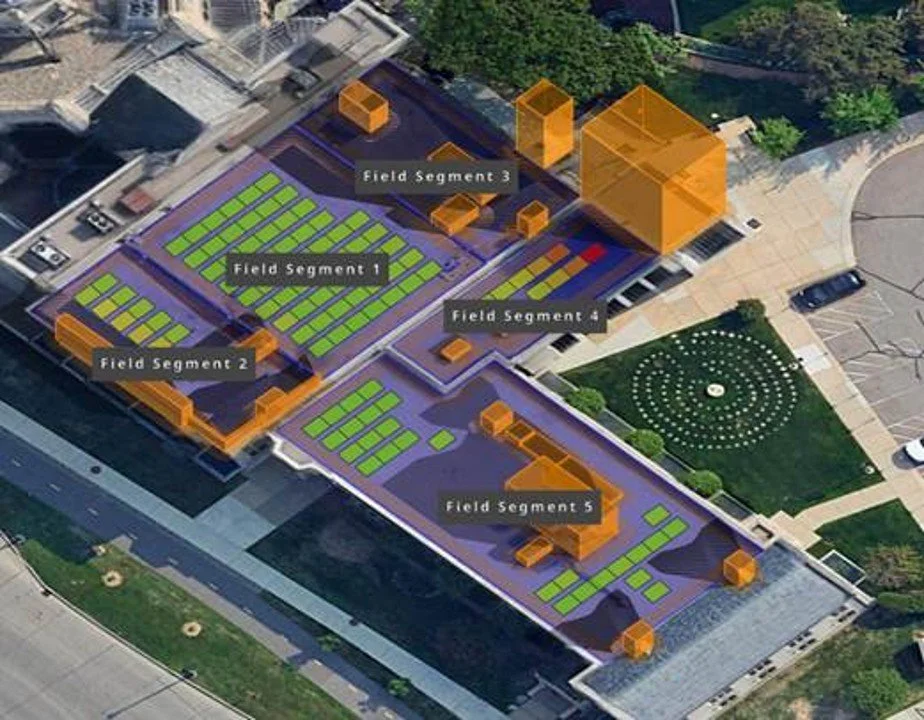 An aerial view of a large rooftop urban farm divided into five field segments with marked areas, several orange structures, green planting beds, and surrounding sidewalks and parking.