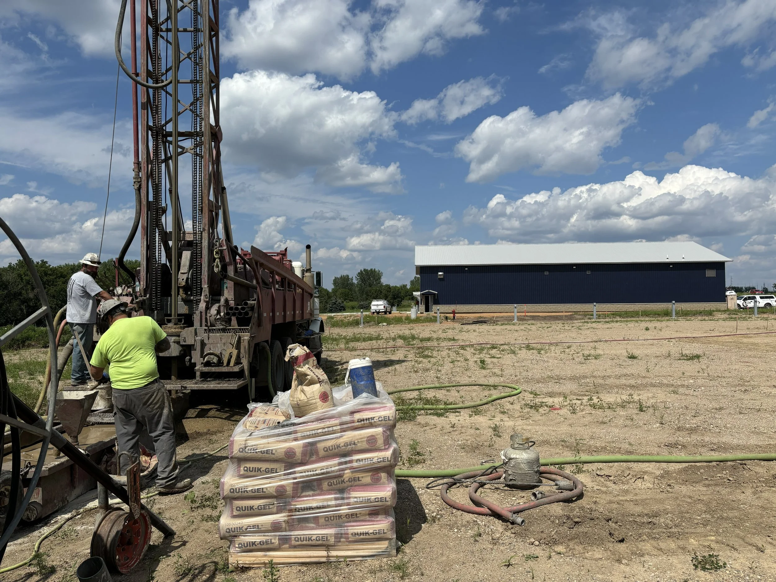 Construction site with workers operating a large drilling machine, stacks of QUIK-GEL bags in the foreground, and a large blue building in the background under a partly cloudy sky.